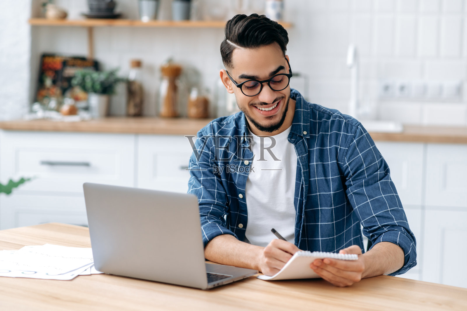快乐有魅力的阿拉伯人或印度人戴着眼镜，坐在家里的桌子上，在一个在线网络研讨会或讲座期间在笔记本电脑上做笔记，使用笔记本电脑，微笑，获得知识照片摄影图片