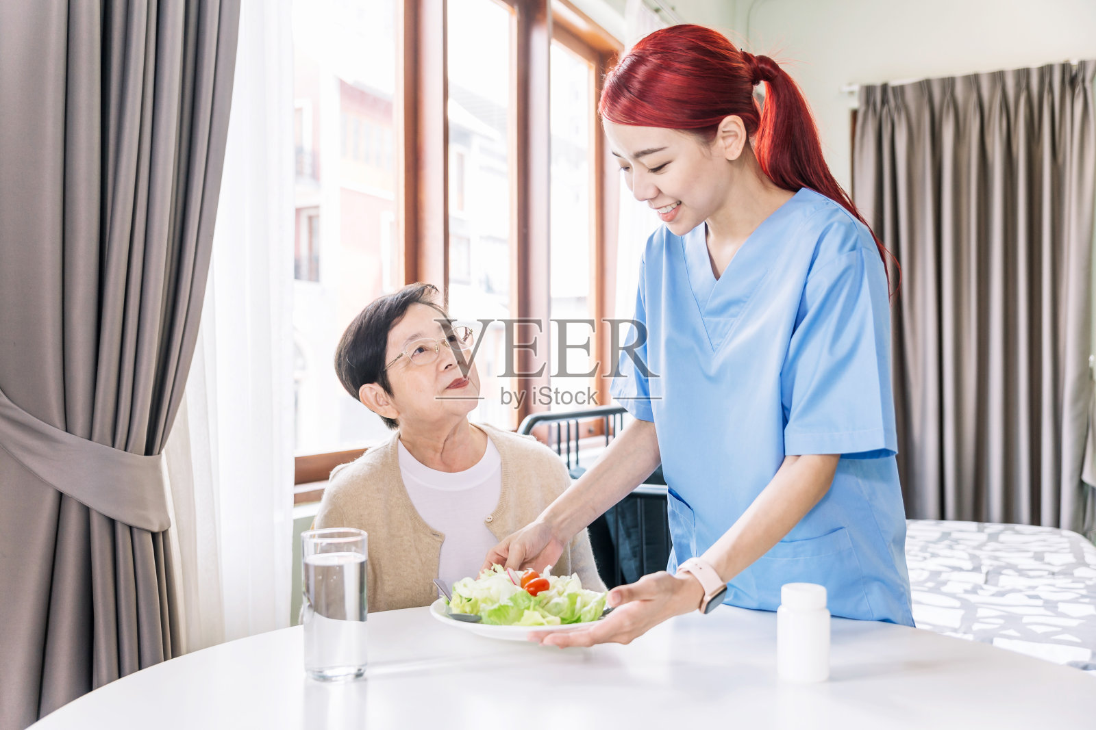 助理亚洲妇女照顾给予照顾老年妇女成人在家里吃沙拉餐。护理人员拜访家庭。家庭保健和疗养院概念。照片摄影图片