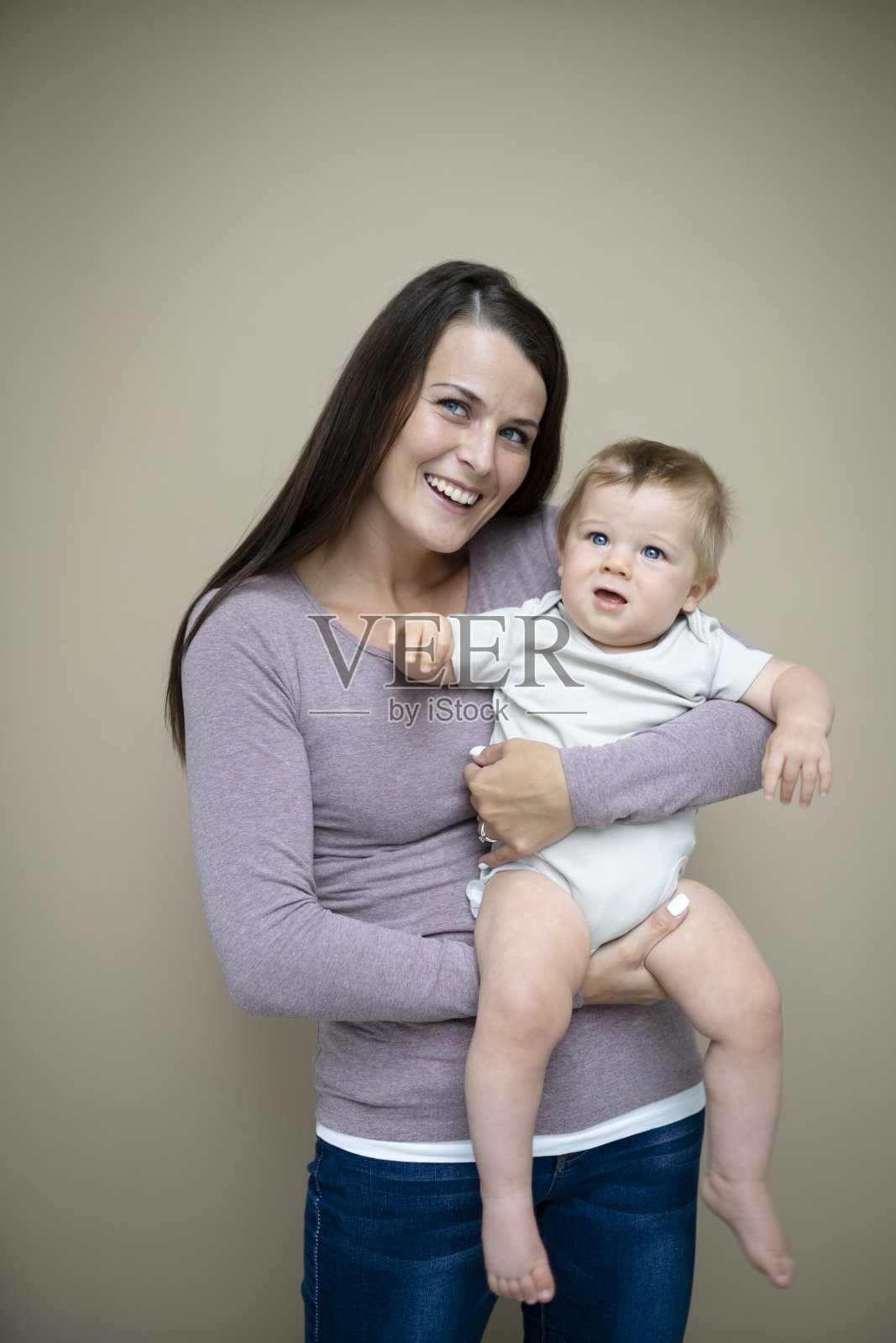 漂亮的年轻妈妈，深色头发，紫色上衣，抱着她7个月大的宝宝，站在棕色背景前照片摄影图片