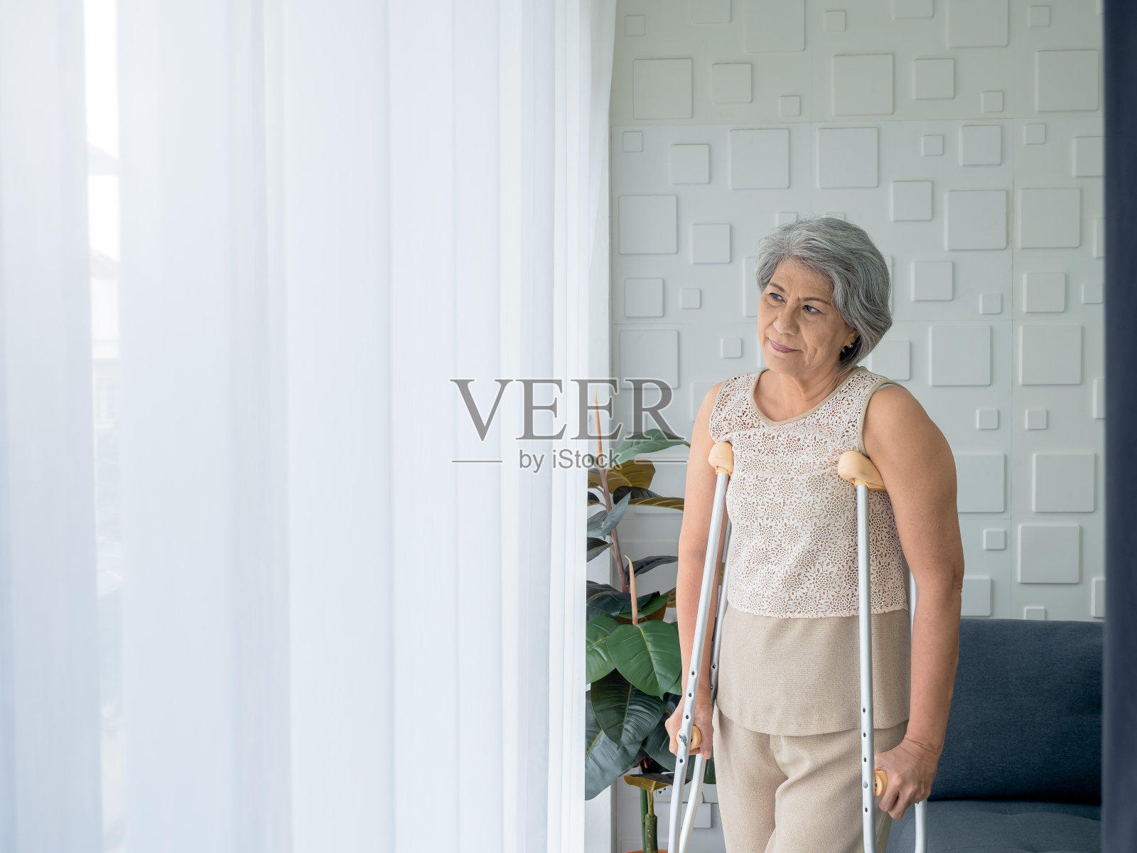 亚洲老年妇女白头发站在步行拐杖附近的窗帘看窗外的复印空间。老妇病人使用拐杖助行器。身体健康，医疗保健观念强。照片摄影图片