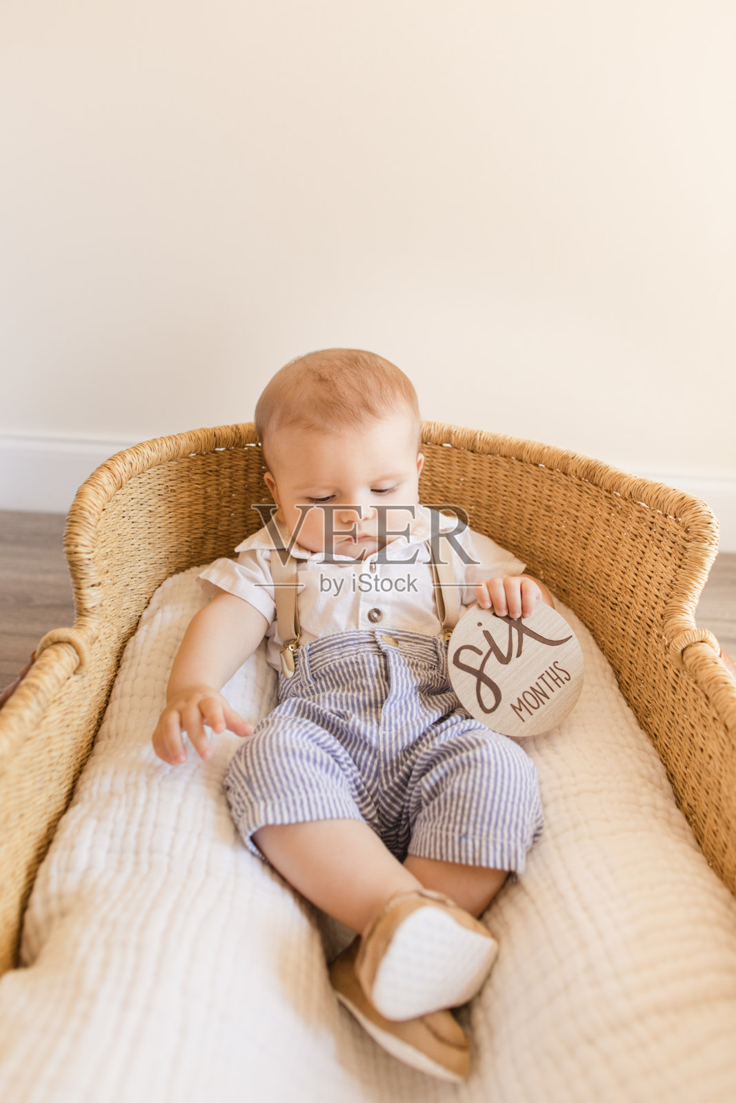 6个月大的小男孩穿着棕色背带的白衬衫，蓝白条纹长裤和棕色乐福鞋，躺在海草摩西篮子里，篮子上有一个木制的圆圈，上面写着“六个月”照片摄影图片