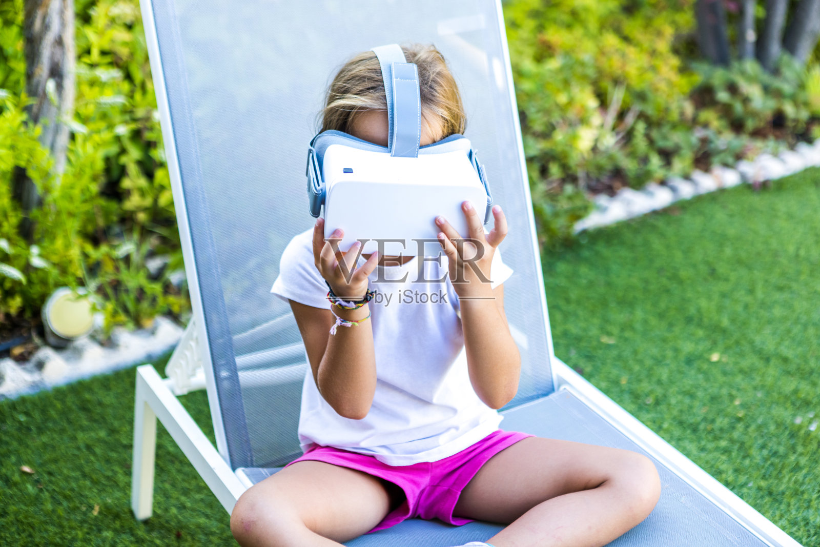 带着虚拟现实眼镜的小女孩，用她的手拿着它们，从上面，坐在她家花园的阳光躺椅上。元宇宙，VR，游戏，数字和模拟概念。照片摄影图片