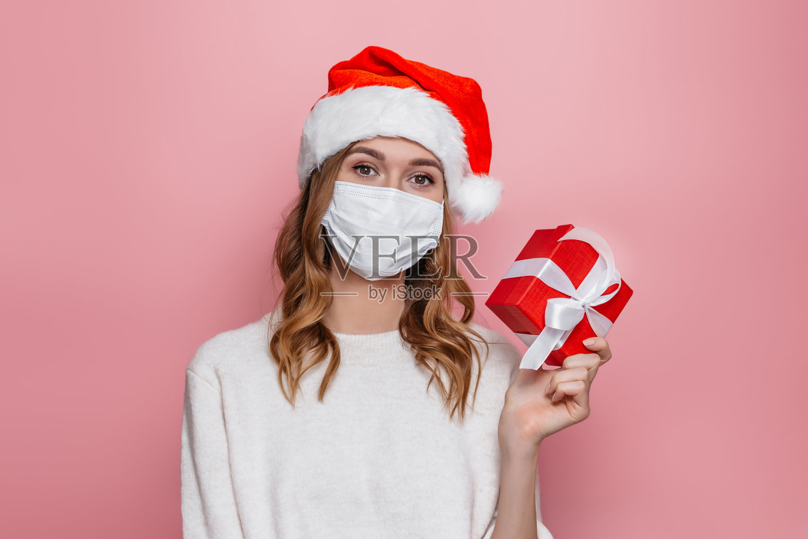 戴着医用口罩和圣诞帽的年轻女子手持红色礼盒，隔离在粉色的工作室背景上。新冠病毒检疫期间的新年礼物概念照片摄影图片