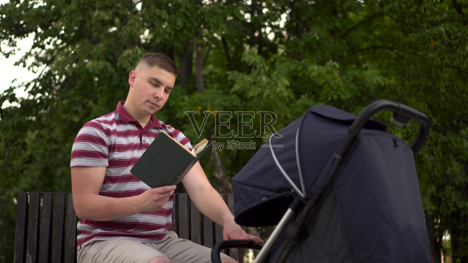 一个年轻人推着婴儿车坐在公园里看书。爸爸坐在公园里的长凳上摇晃婴儿车。照片摄影图片