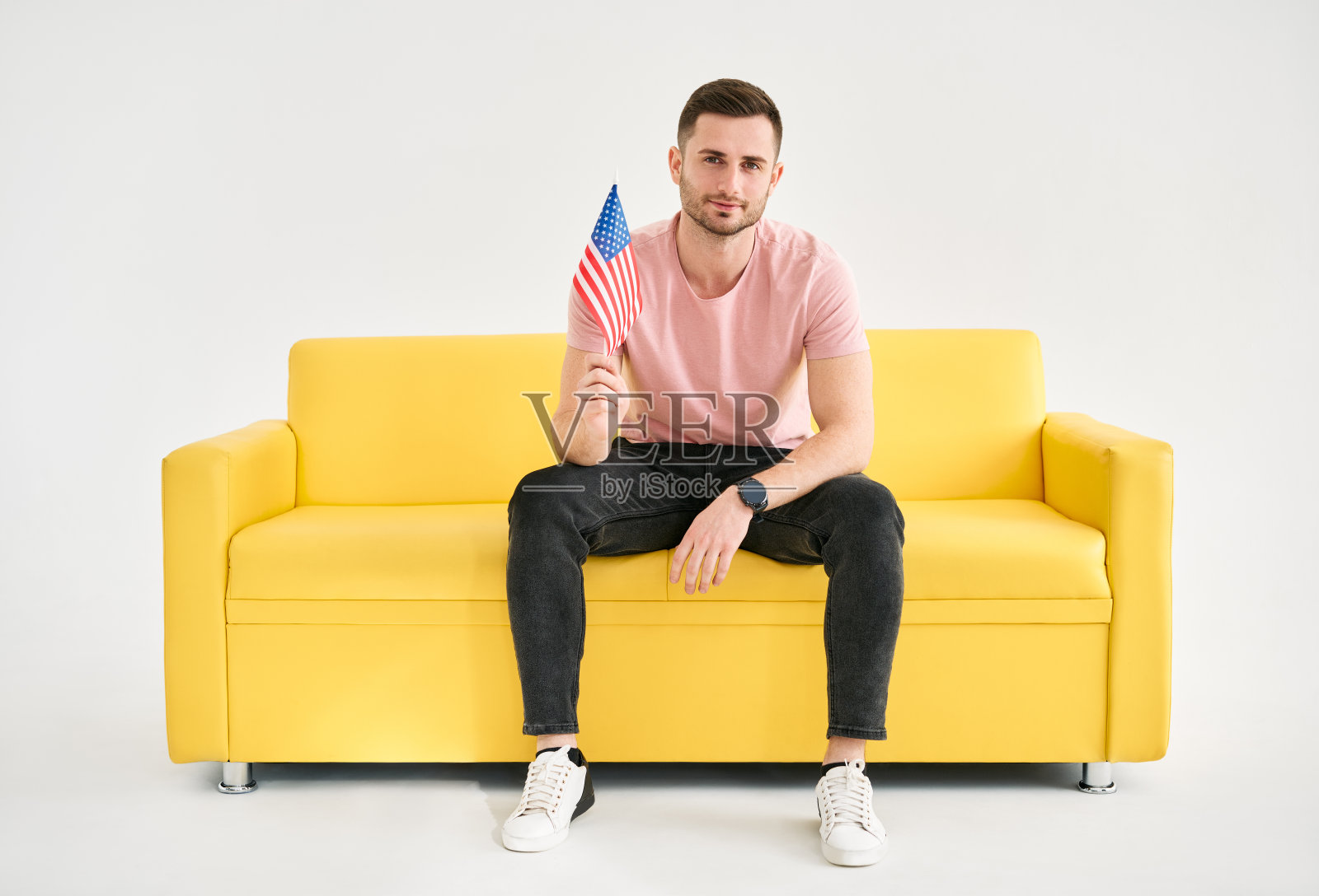 年轻快乐的人与美国国旗在他的手坐在明亮的沙发覆盖白色背景照片摄影图片