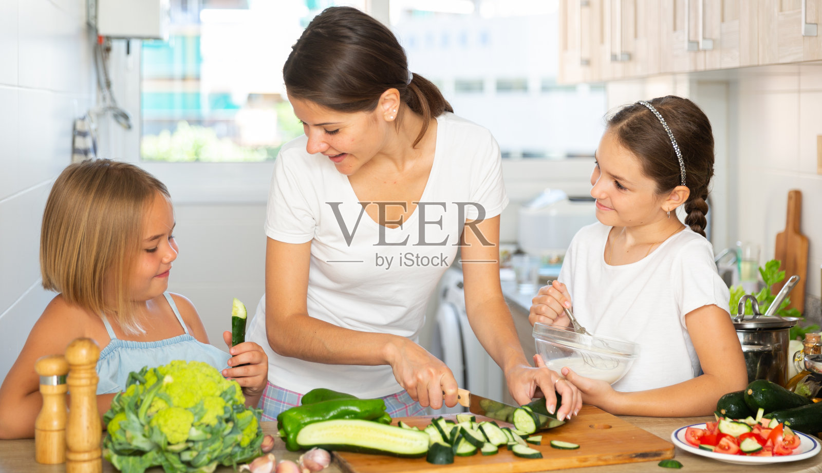 快乐的女人带着两个小女儿在厨房里准备蔬菜沙拉照片摄影图片