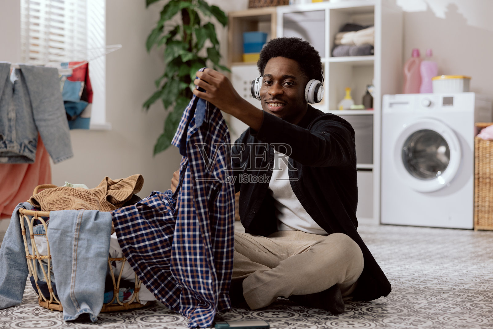 画像的一个微笑的年轻男子折叠干净的衣服，分类衣物之前把它放在洗衣机，一个学生坐在浴室地板上听音乐的无线耳机照片摄影图片