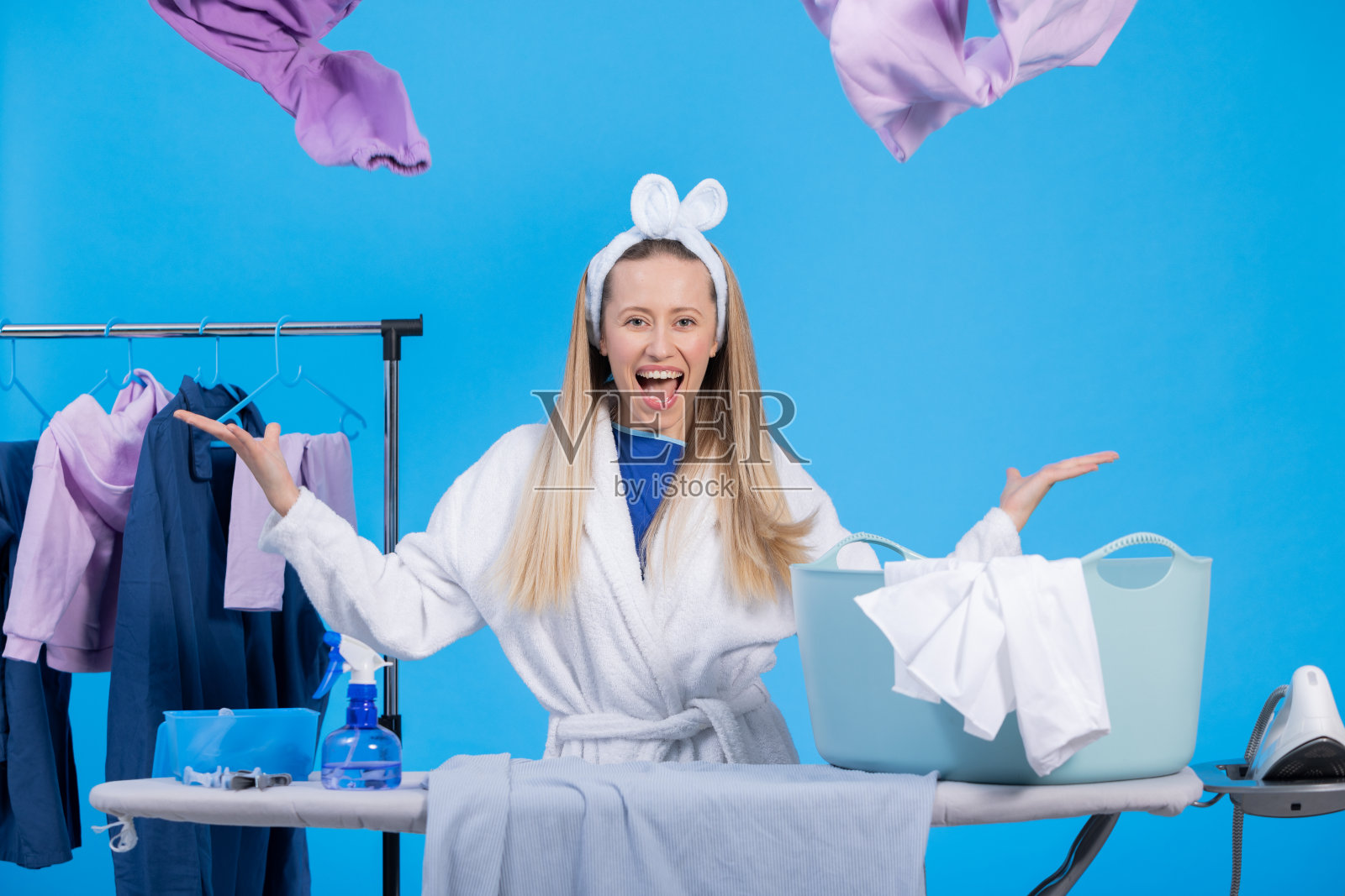 微笑的女孩从碗里扔出需要折叠、分类、熨烫的衣服。家庭主妇喜欢在家里做家务。在工作室蓝色背景的肖像照片摄影图片