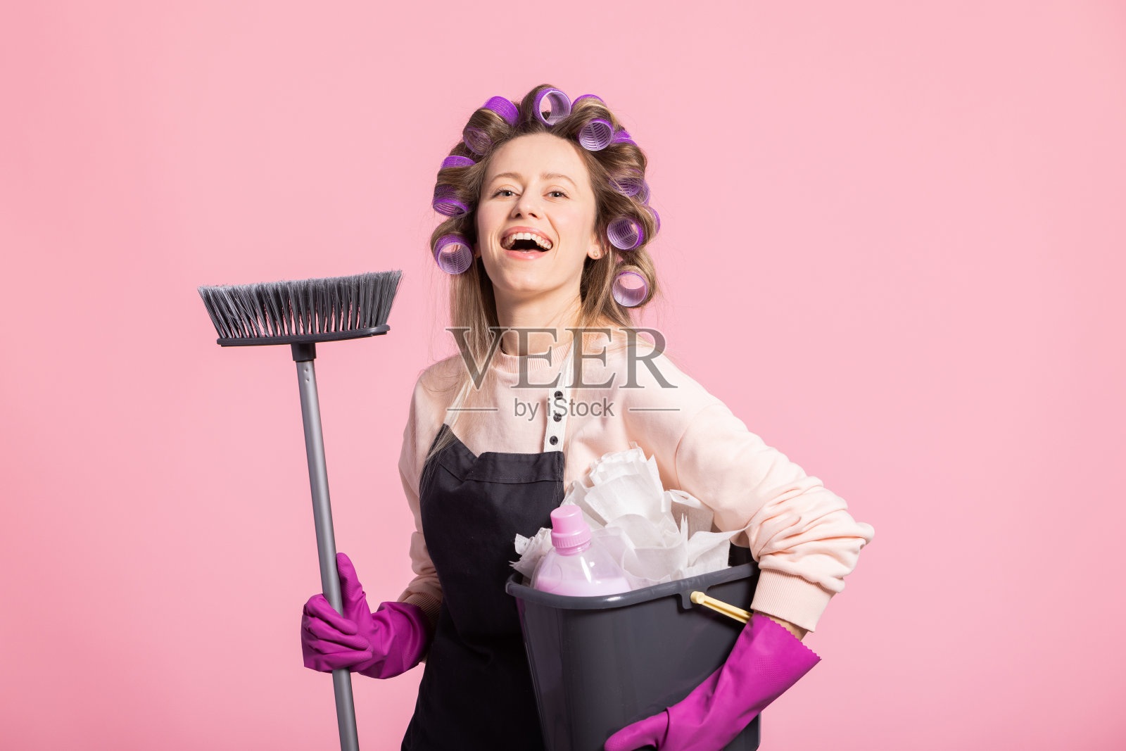 一个梳着卷发的女孩笑眯眯地看着镜头，心满意足地拿着扫帚和拖把桶。女人准备拖地的公寓，粉红色的工作室背景照片摄影图片