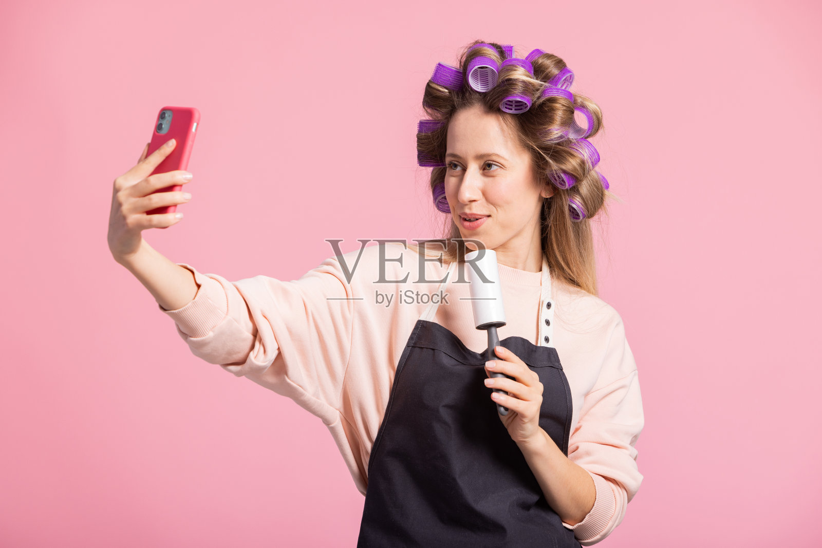 粉色背景上的女人肖像。头戴卷发卷的女孩微笑着对着洗衣服卷唱歌录视频，对着相机打电话照片摄影图片