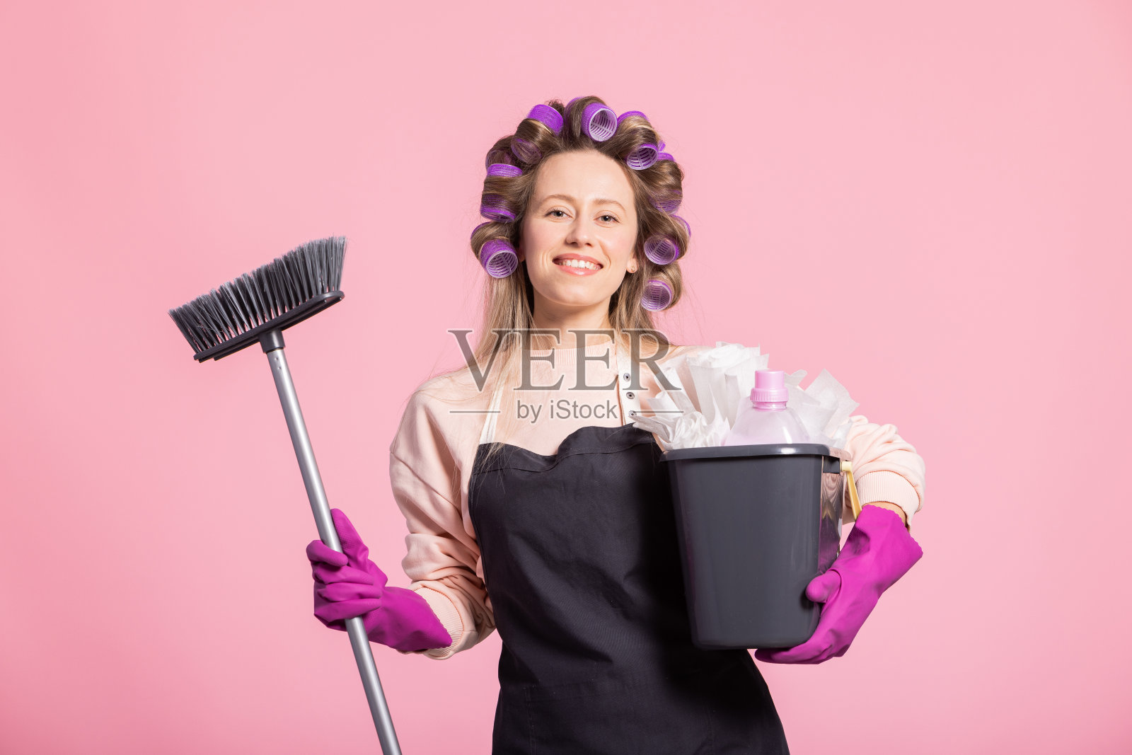 漂亮的家庭主妇的画像好心情的妻子拿着清洁设备明亮的粉红色背景女人戴着乳胶手套粉红色的围裙和头上的滚筒照片摄影图片