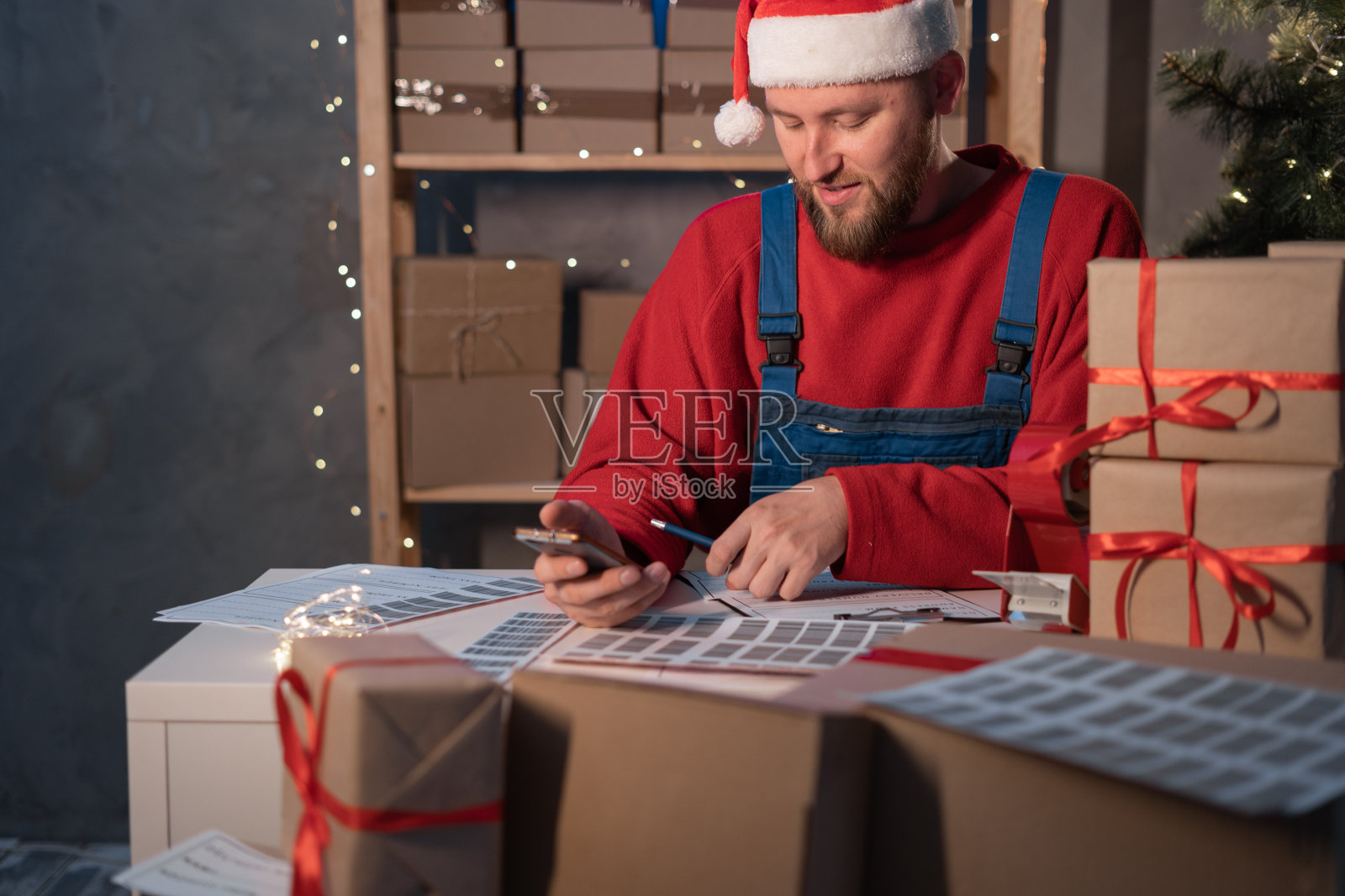 圣诞老人商人坐在他的办公桌前，用他的手机扫描一个快递箱。圣诞节，一名男子在网上销售商品，用他的智能手机扫描条形码。照片摄影图片