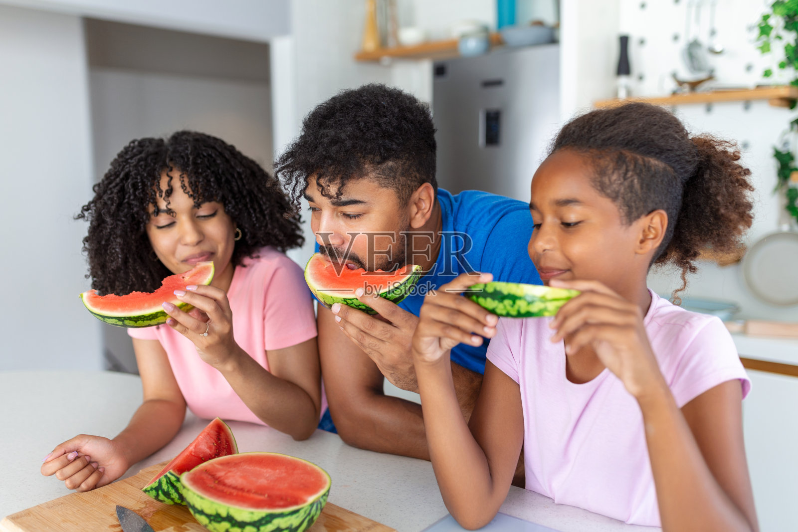 年轻的一家人吃西瓜，玩得很开心。混血家庭在厨房里一起吃一片西瓜。照片摄影图片