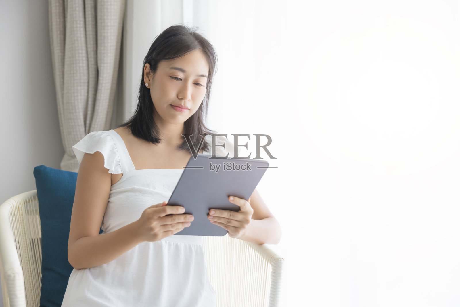 可爱的快乐的亚洲女人穿着白色的裙子，在奶油色的沙发上玩他的平板电脑。看手机屏幕照片摄影图片