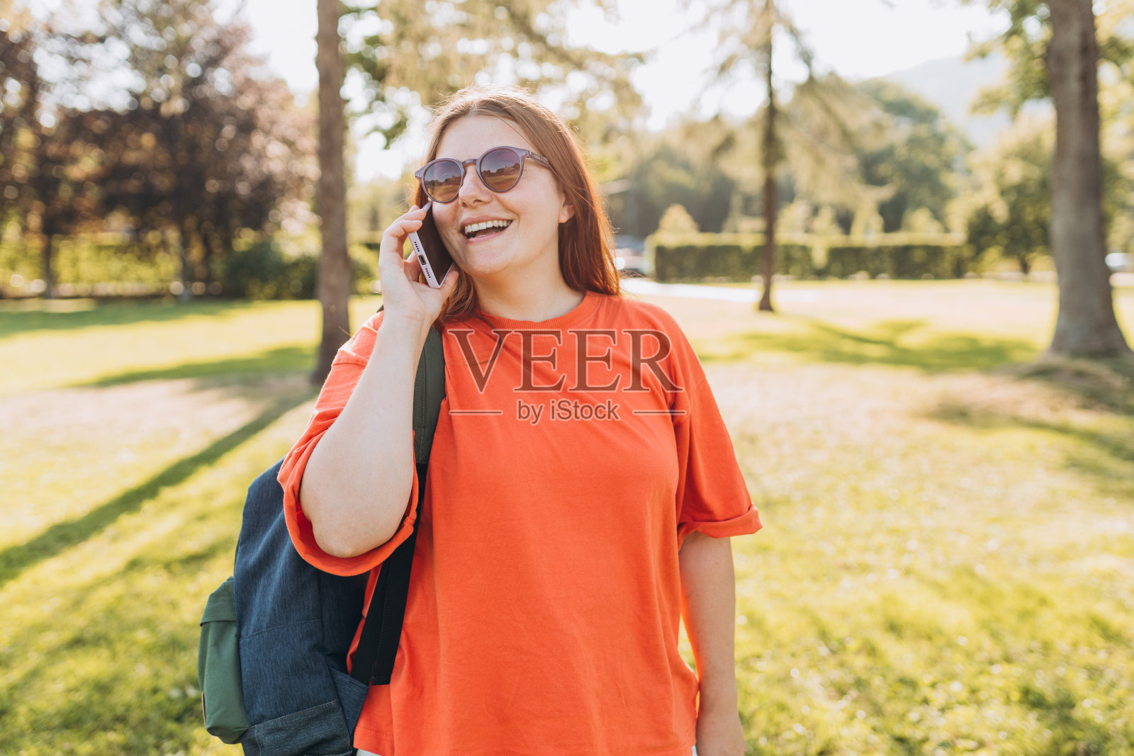 在自然背景下，一个微笑着打电话的美丽女人的肖像。夏天，背着双肩包的快乐女子正在户外公园里使用智能手机。旅行者,阳光灿烂的日子照片摄影图片