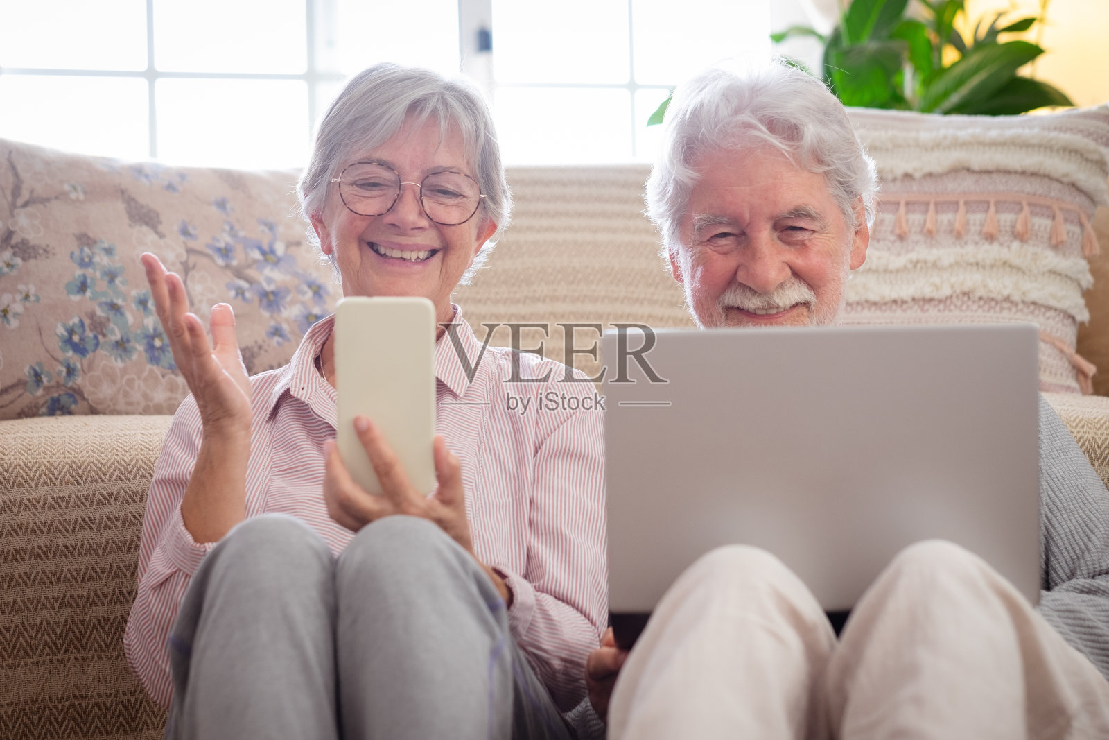 微笑愉快的老夫妇在家放松坐在地板上使用数字设备。丈夫用笔记本电脑，妻子用手机视频通话。照片摄影图片