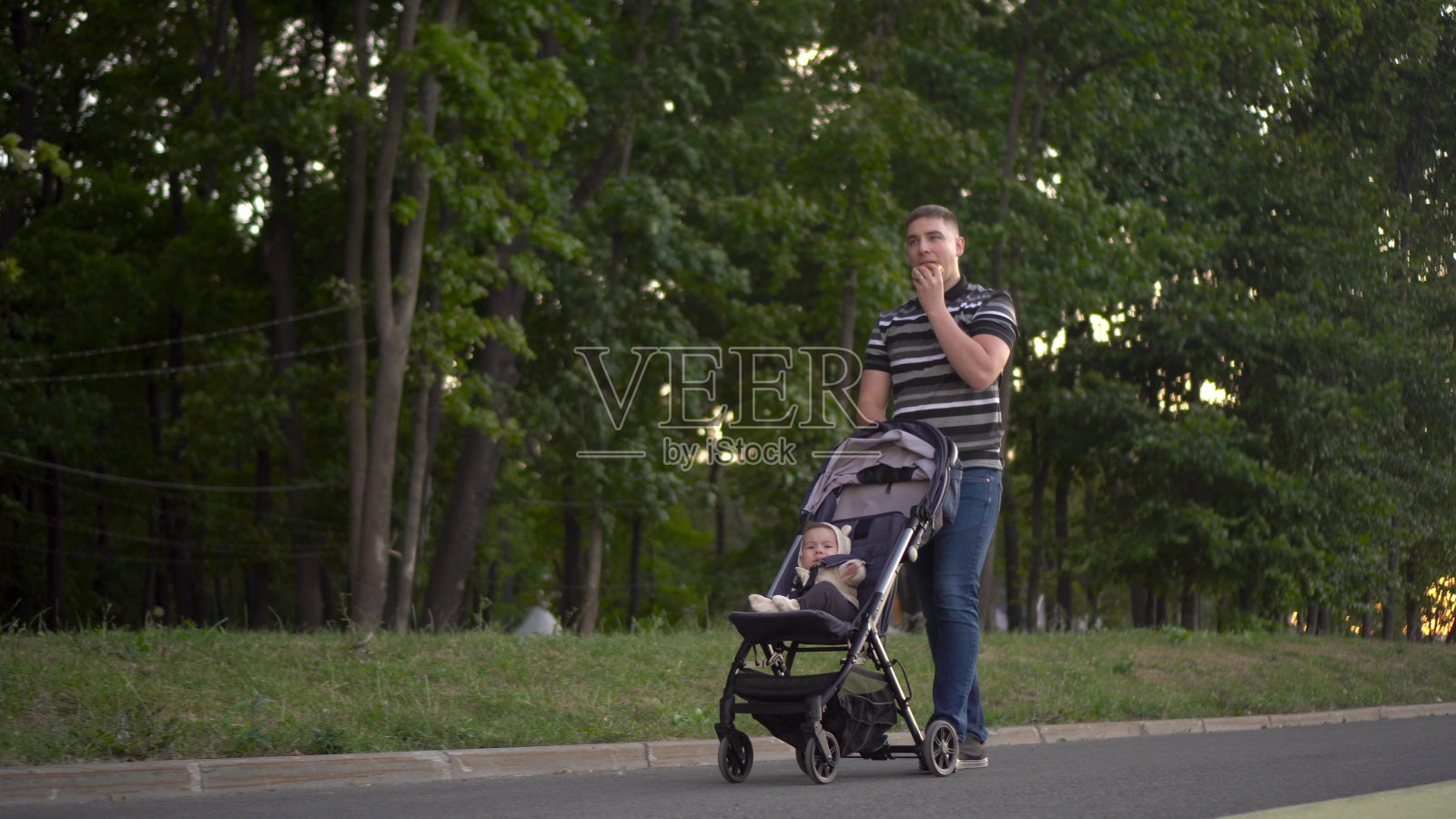 一个男人在公园里滚着一个婴儿，吃着一个汉堡。年轻的父亲带着儿子散步。照片摄影图片
