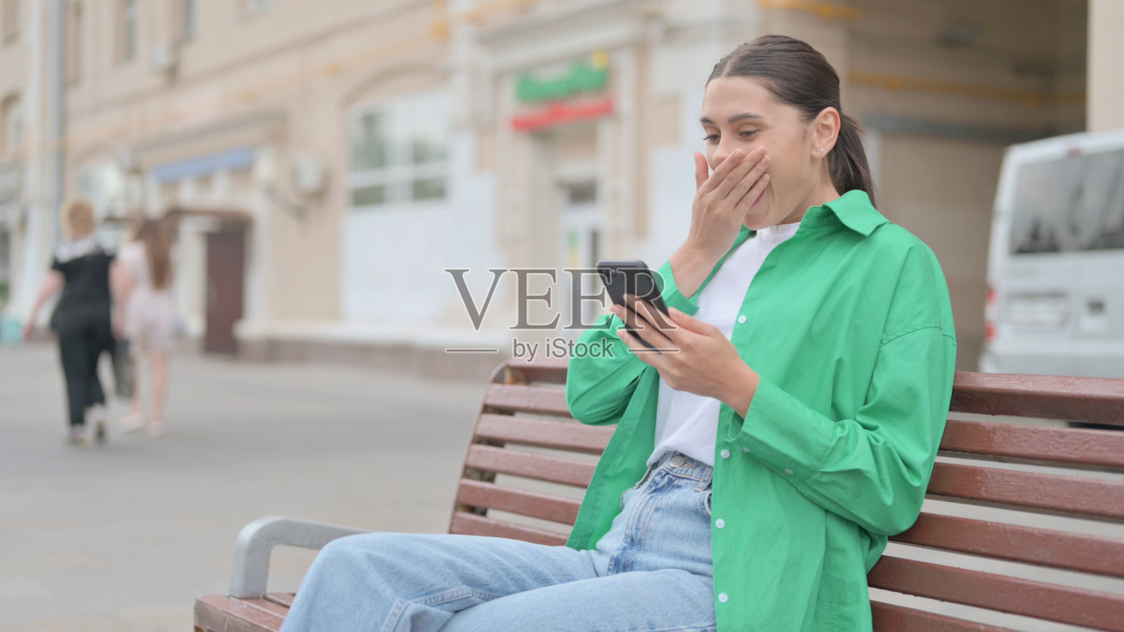 西班牙妇女坐在户外长凳上用智能手机庆祝网上成功照片摄影图片