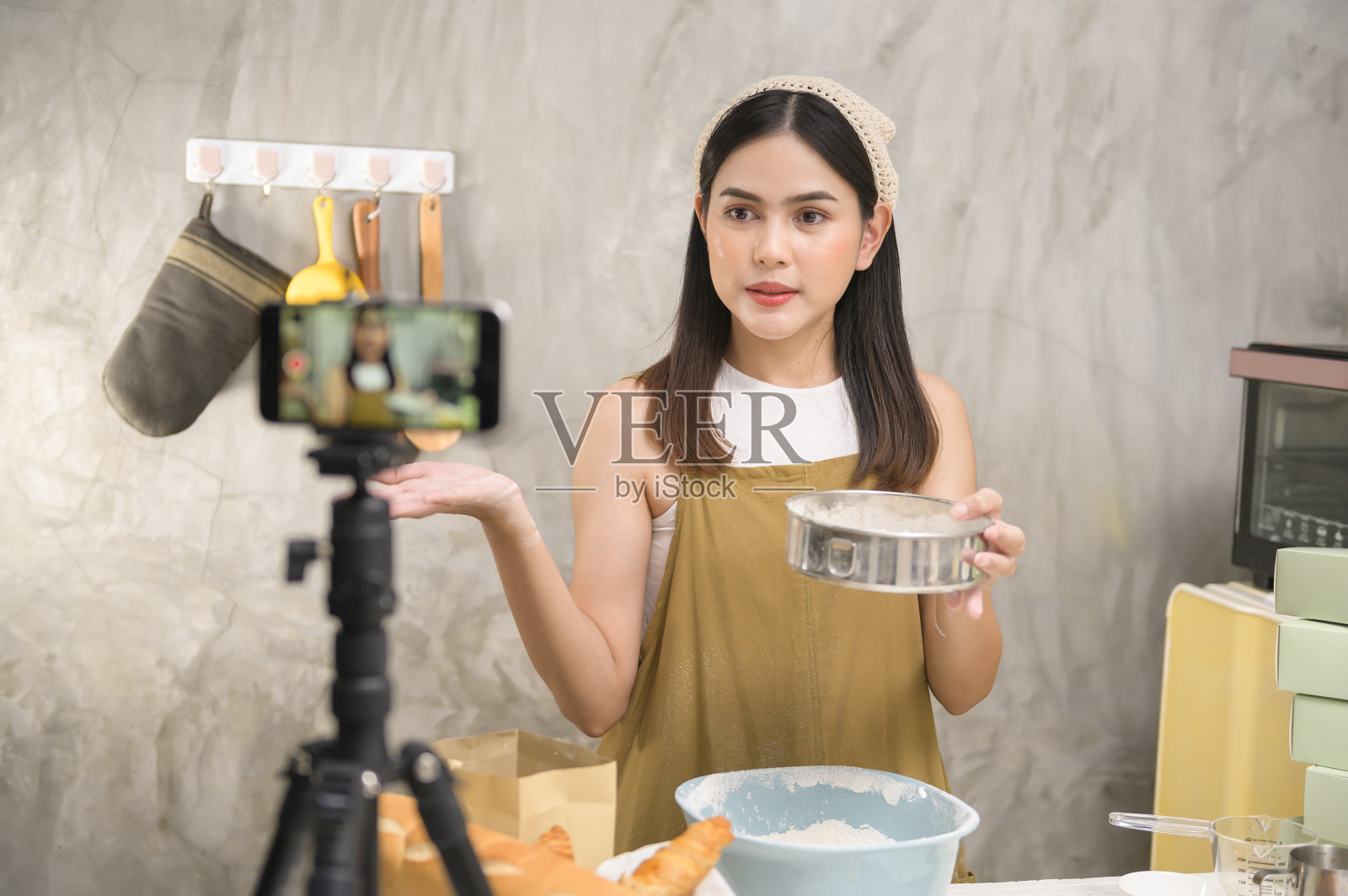 美丽的女人正在做面包，同时直播或在社交媒体上录制视频在她的房子，咖啡店的商业概念。照片摄影图片