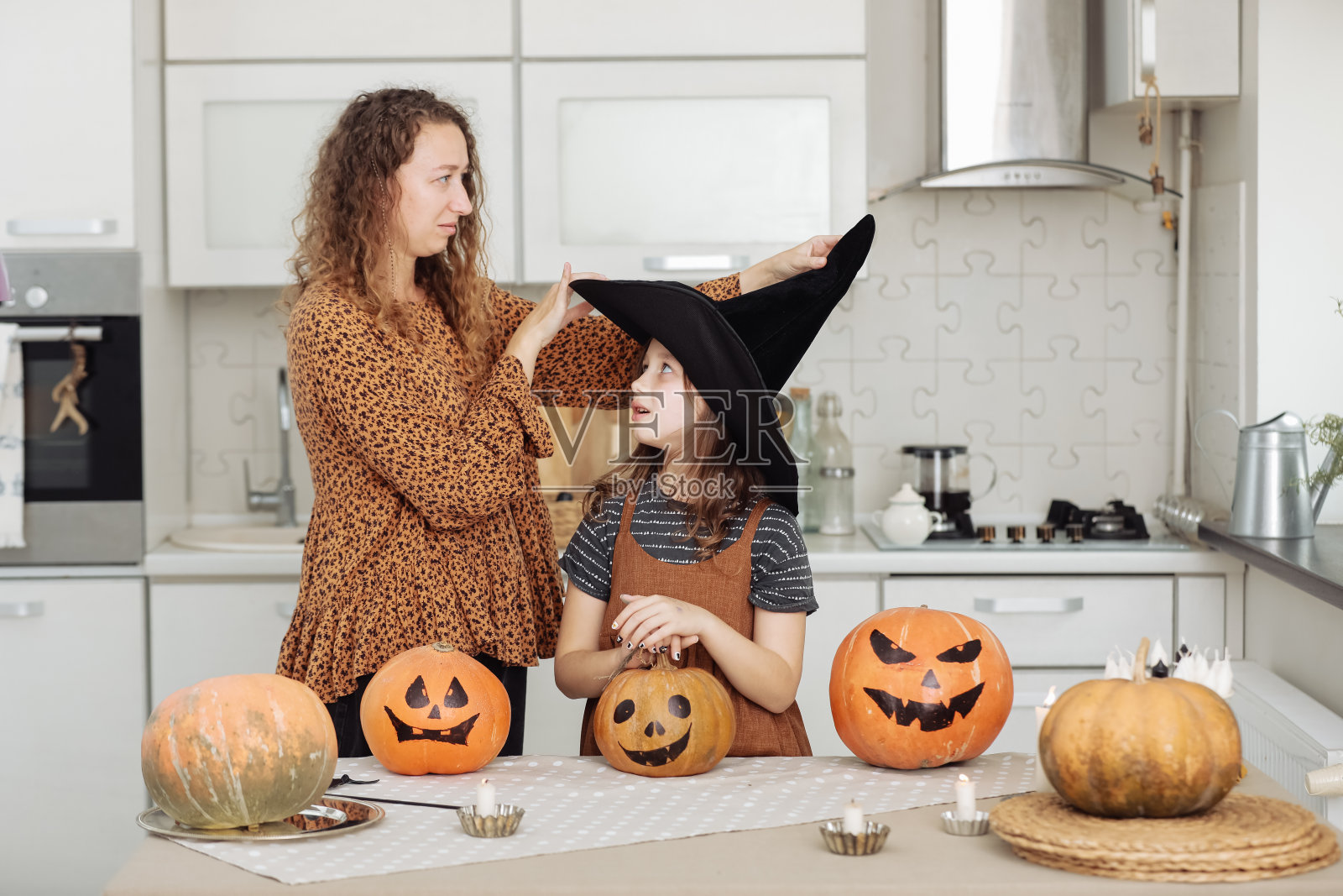 幸福的家庭:妈妈和女儿庆祝万圣节。房间里穿着女巫狂欢节服装的快乐孩子。快乐的孩子和父母在家里玩南瓜和黑女巫帽。照片摄影图片