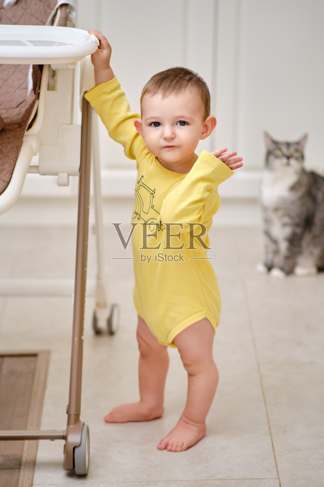 一岁的小男孩抱着一张儿童高脚椅站在厨房里照片摄影图片