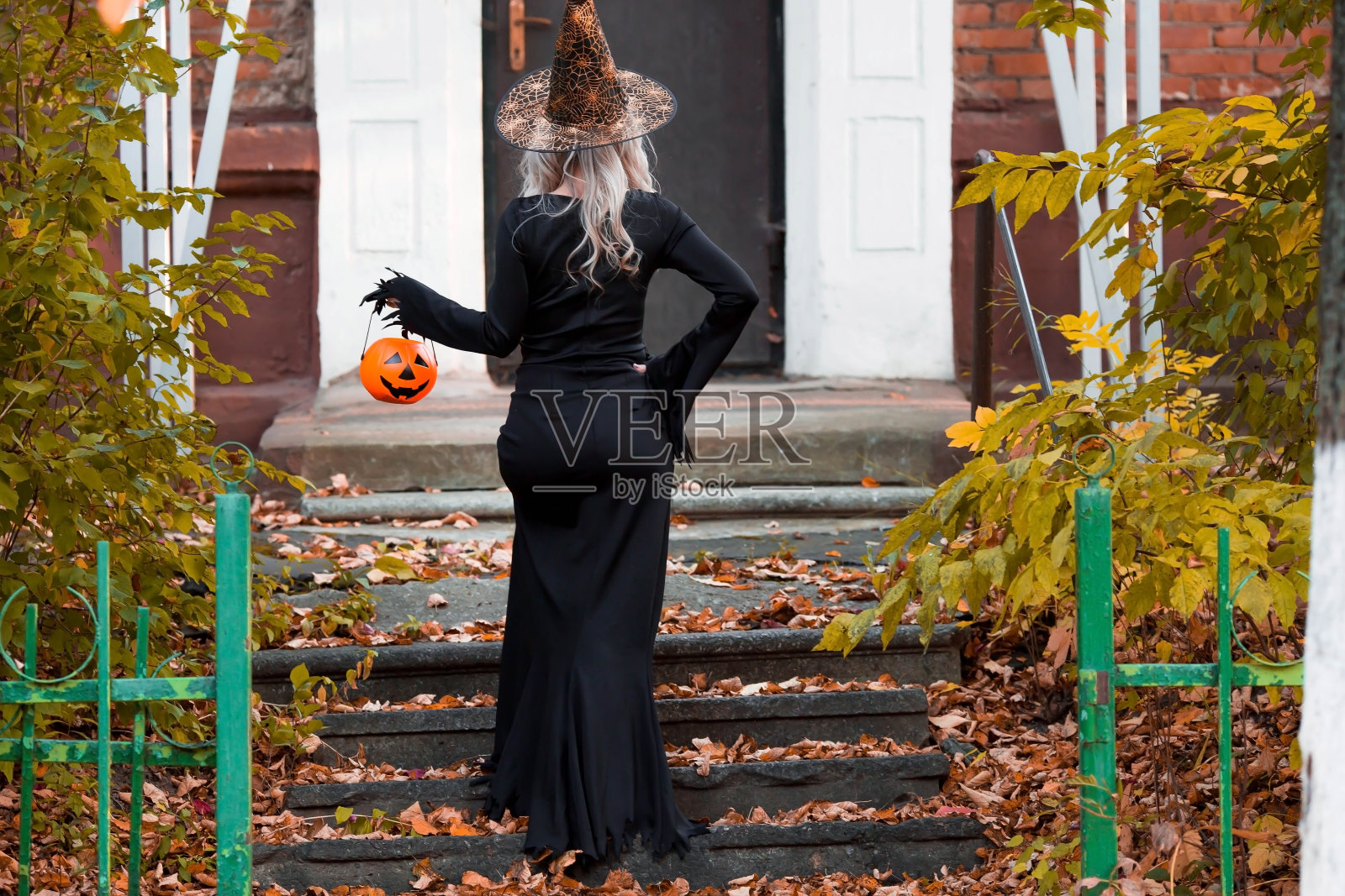 万圣节快乐。一个穿着黑裙子、戴着女巫帽、手里拿着南瓜桶的女人。在户外穿着嘉年华服装的有趣的人们。照片摄影图片