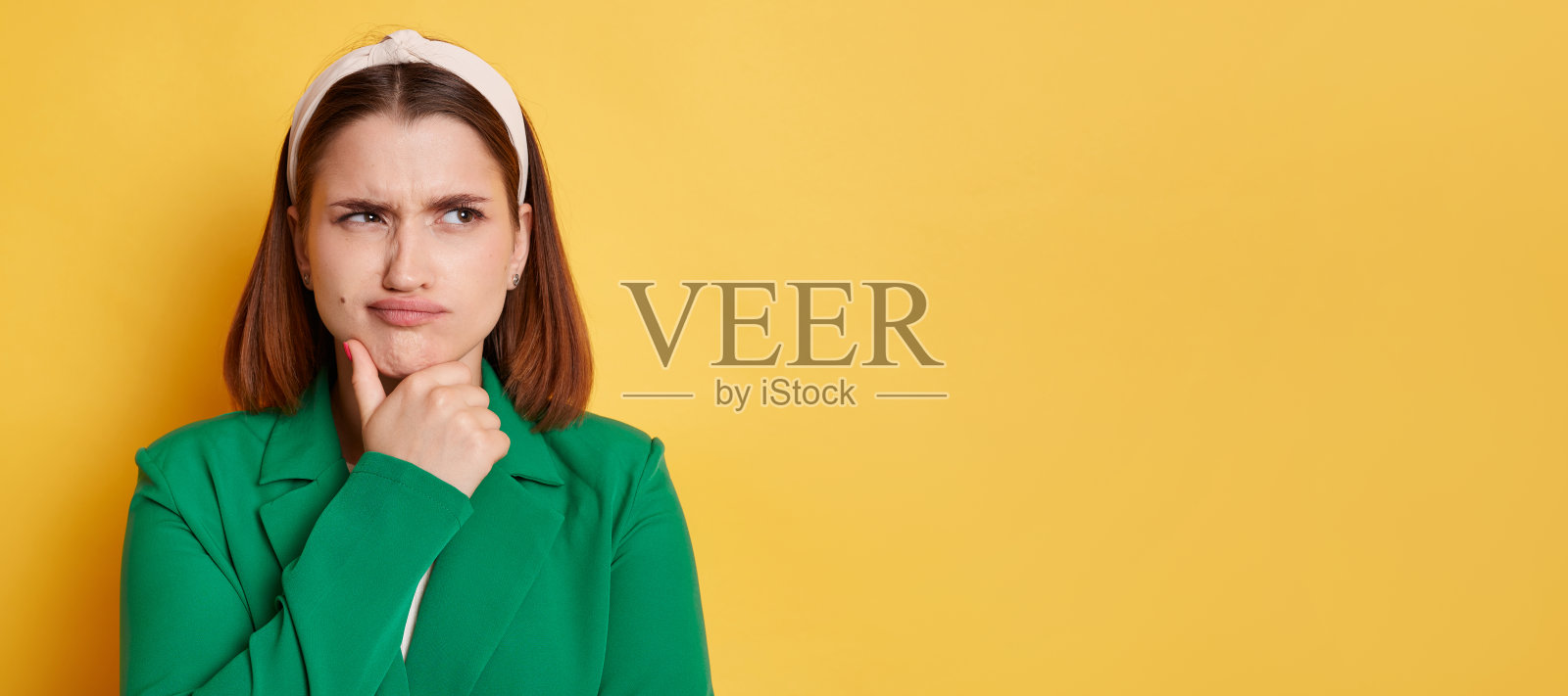 肖像沉思的妇女穿着绿色夹克和发带摆姿势孤立在黄色背景，女性抱着下巴和思考，复制空间为广告。照片摄影图片