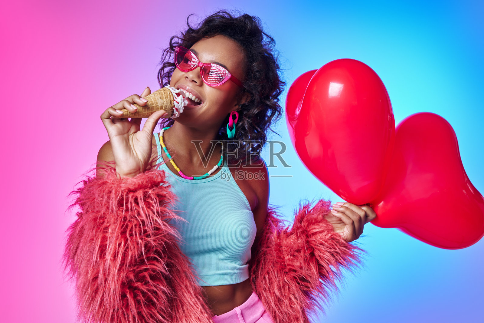快乐的非洲妇女穿着蓬松的夹克，拿着气球，在五颜六色的背景下享受冰淇淋照片摄影图片