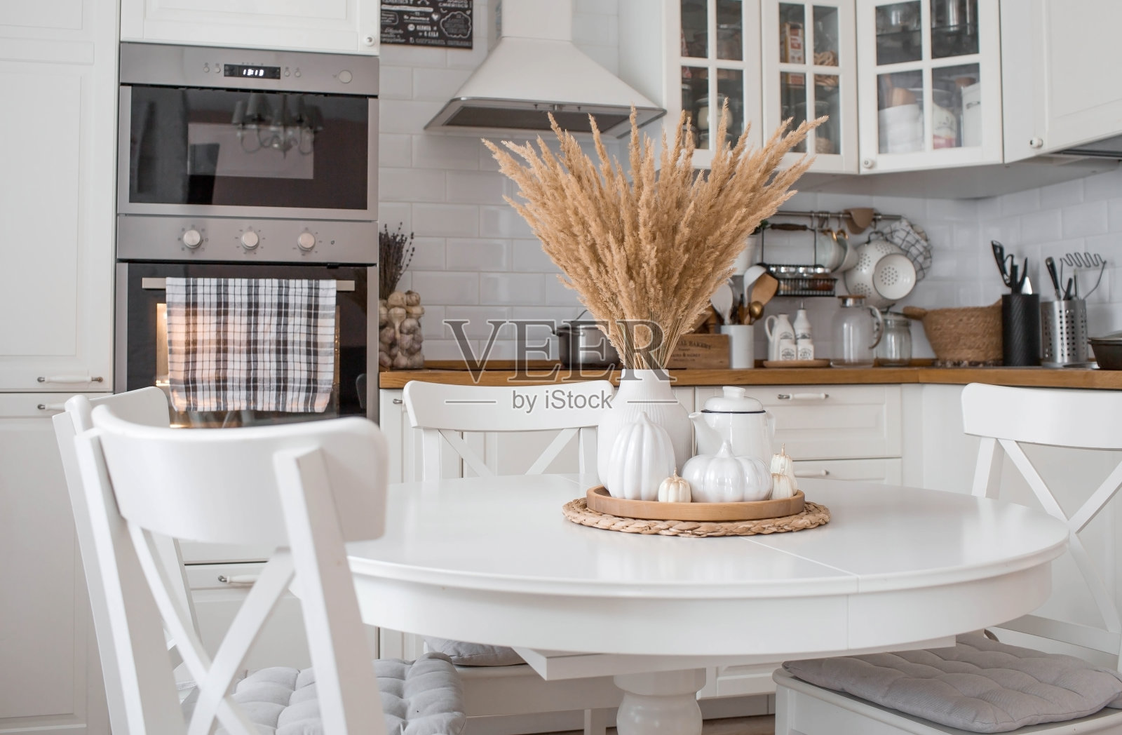 静物。在斯堪的纳维亚风格的家庭厨房里，白色的桌子上放着花瓶里的干潘帕斯草、白色的陶瓷南瓜、茶壶、杯子和南瓜形状的蜡烛。舒适的秋天的概念。照片摄影图片