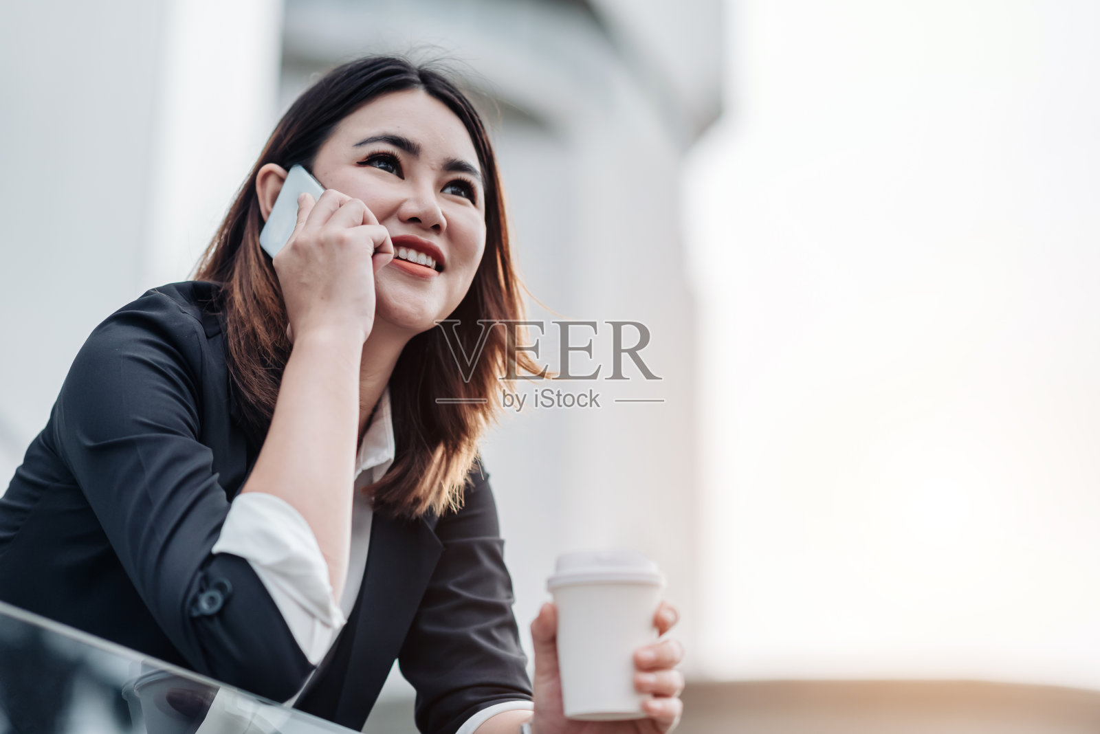 年轻的亚洲女商人在出差前坐在机场打电话。美女乘客拿着手机，微笑着讨论着什么，手里拿着咖啡照片摄影图片
