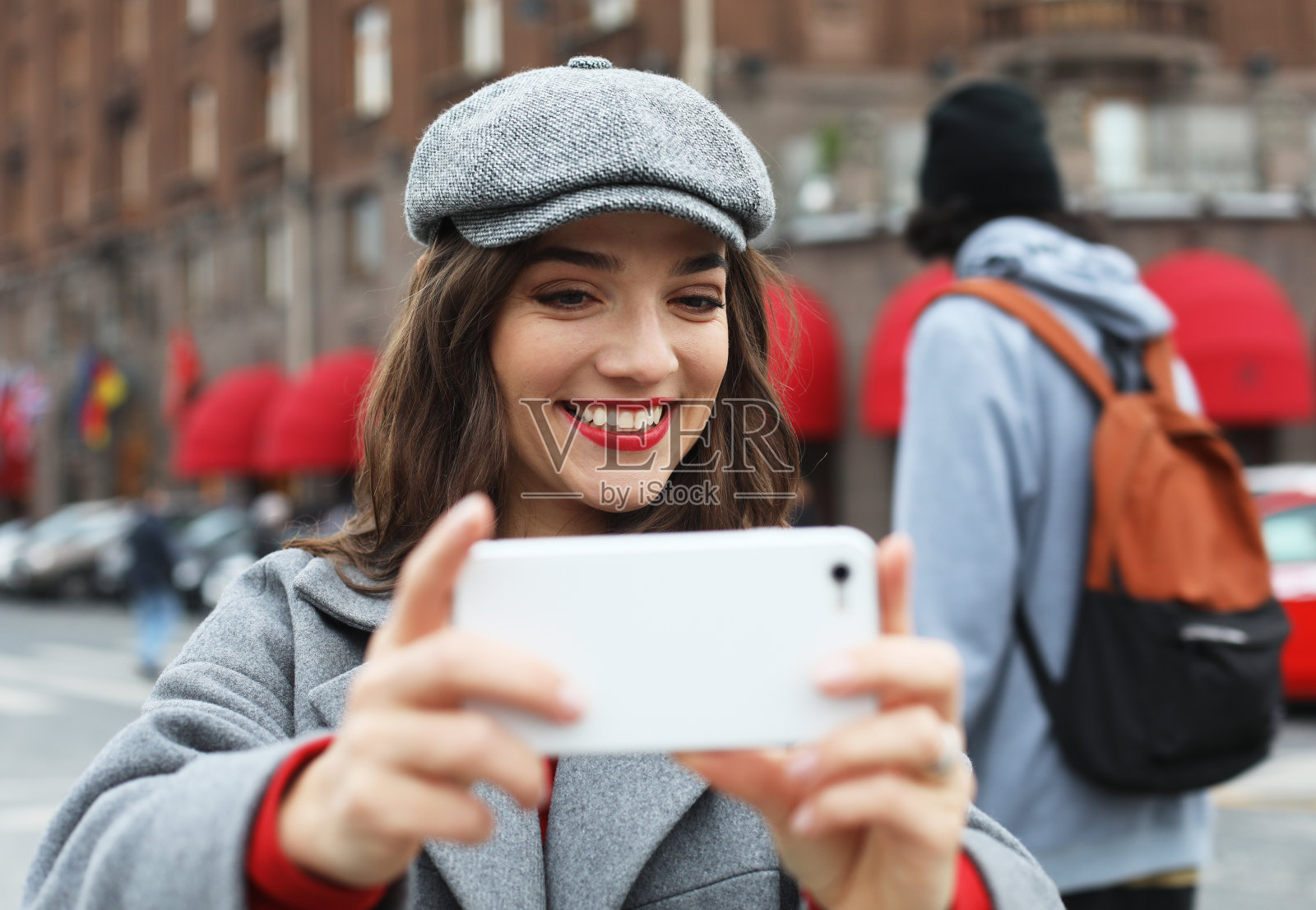 可爱的女人戴着时髦的灰色帽子和外套在走过老建筑时自拍。照片摄影图片