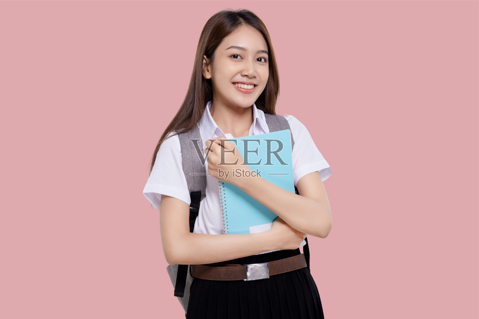 肖像微笑的十几岁的亚裔学生女孩在大学制服举行背包孤立的粉红色背景。照片摄影图片