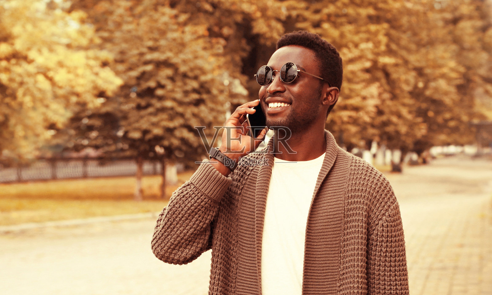 画像快乐微笑的非洲人打电话在智能手机，穿着棕色针织开衫和太阳镜在城市街道上秋天公园的背景照片摄影图片