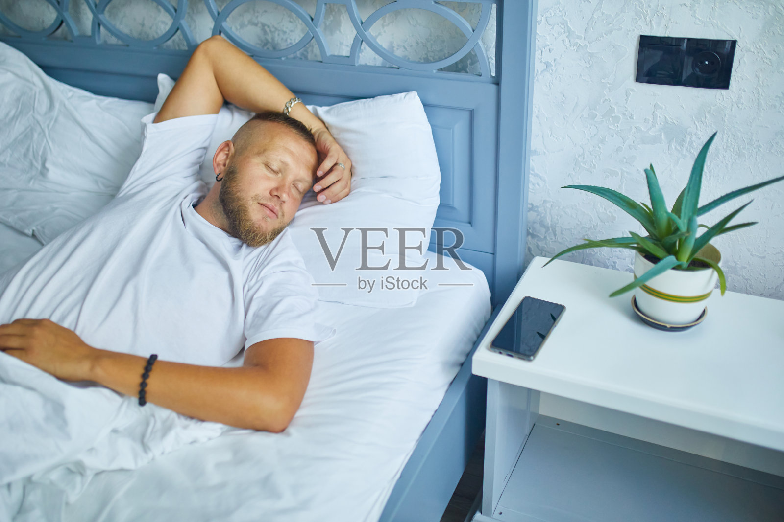 大胡子男人独自睡在一个又大又舒适的床上，白色亚麻布和智能手机闹钟在家里，生活方式，舒适的枕头，床照片摄影图片