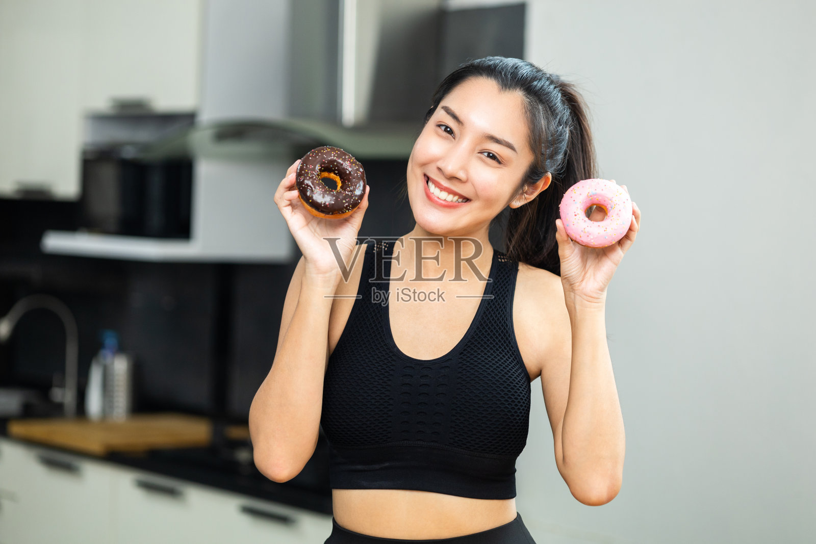 美丽的女性穿着运动服吃甜甜圈甜点。饥饿的年轻女子拿着巧克力甜甜圈不健康食品。食物和甜点概念。照片摄影图片