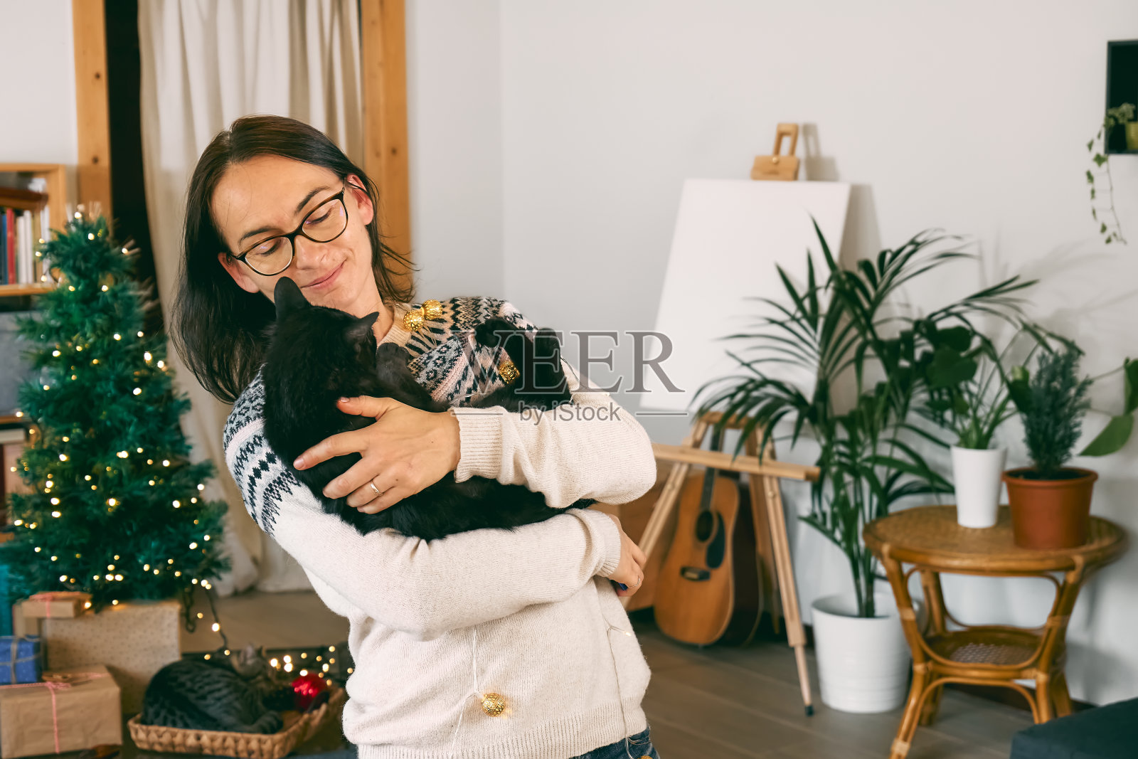 年轻微笑的女人拥抱黑色的家猫在节日圣诞灯在现代房间与礼物和圣诞装饰。快乐的寒假。照片摄影图片