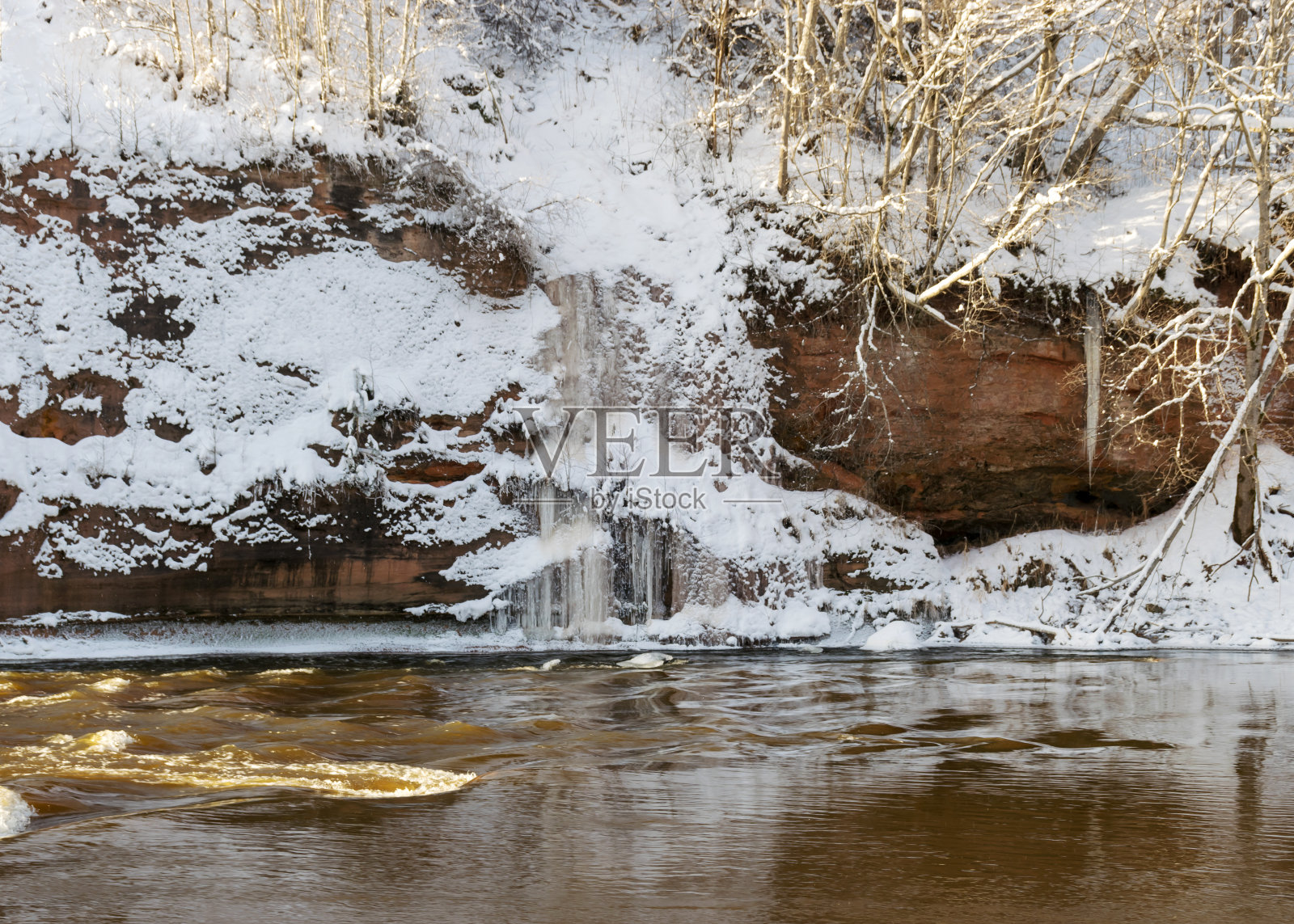 阳光明媚的日子，红色的砂岩峭壁被雪覆盖，悬崖壁上结冰的冰柱，悬崖壁上的冰瀑，冰冻的河流，Gauja, Kuku悬崖，拉脱维亚照片摄影图片