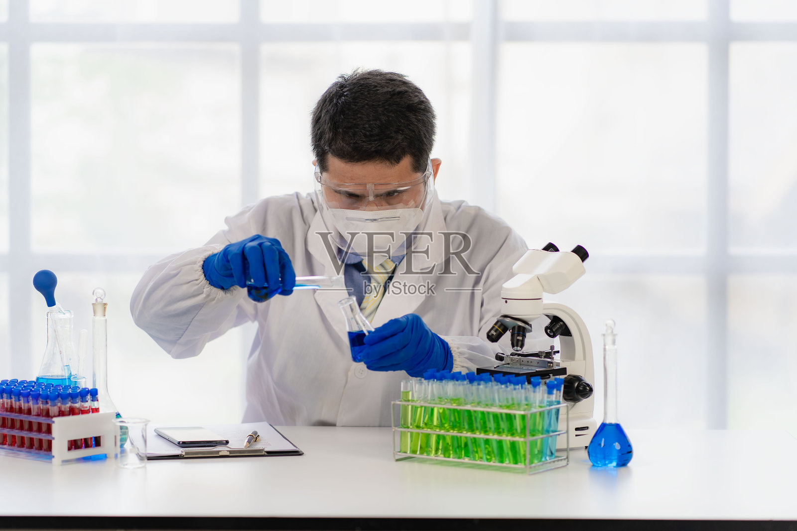 在化学分析实验室，身穿白大褂、戴着蓝色手套的一名男性研究员正在检查装有蓝色液体样品的化学管。照片摄影图片
