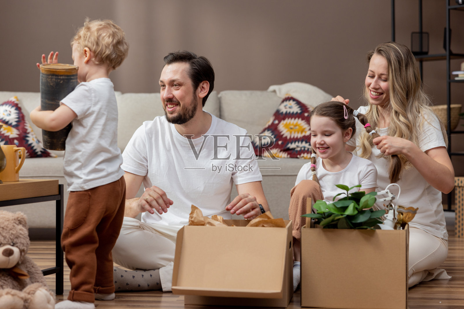 父母带着两个年幼的孩子搬进新公寓，打开纸板箱，拿出东西，可爱的儿子拿着花瓶，妈妈给女儿编辫子照片摄影图片