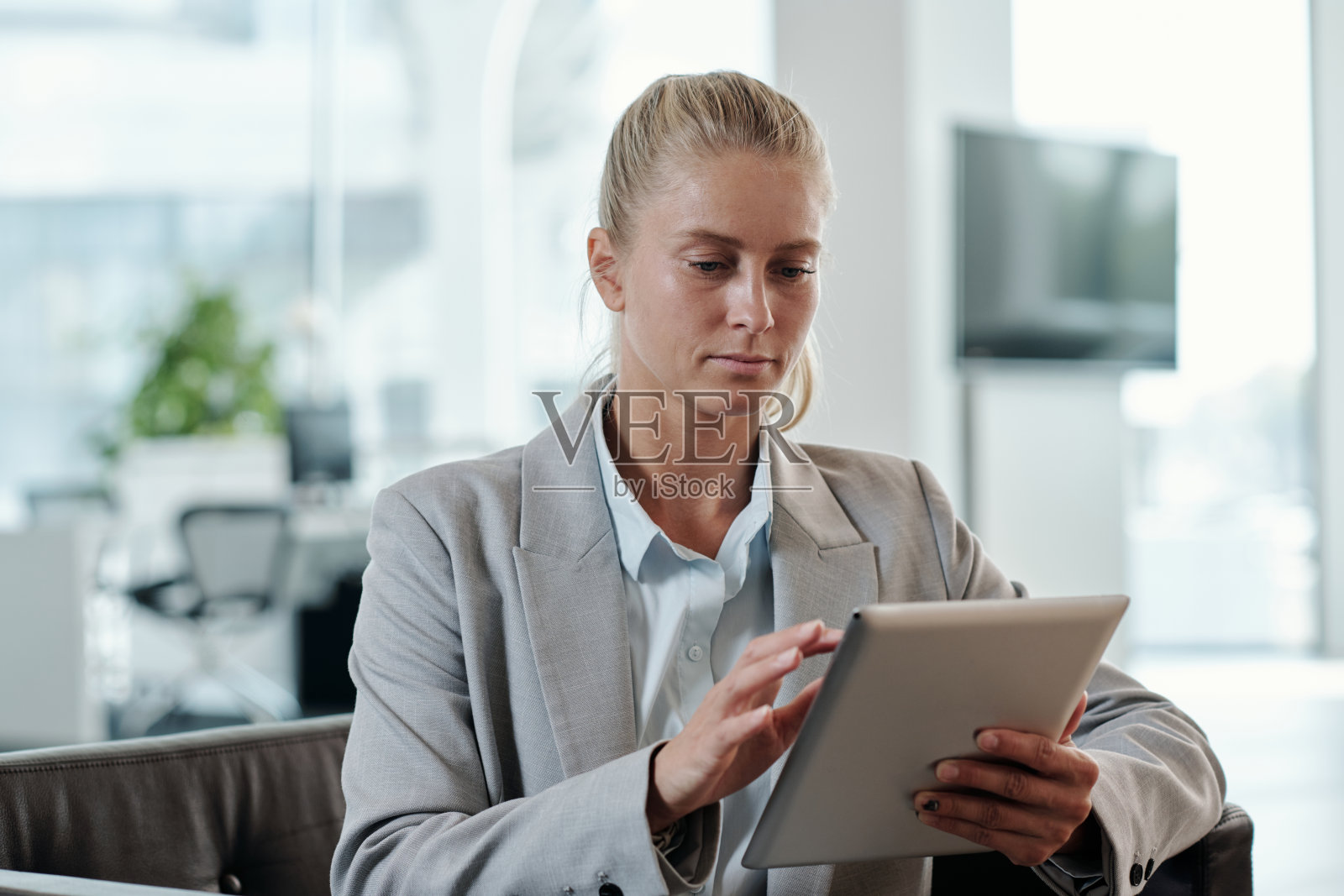 年轻的金发女商人穿着灰色正装在办公室的平板电脑上滚动照片摄影图片