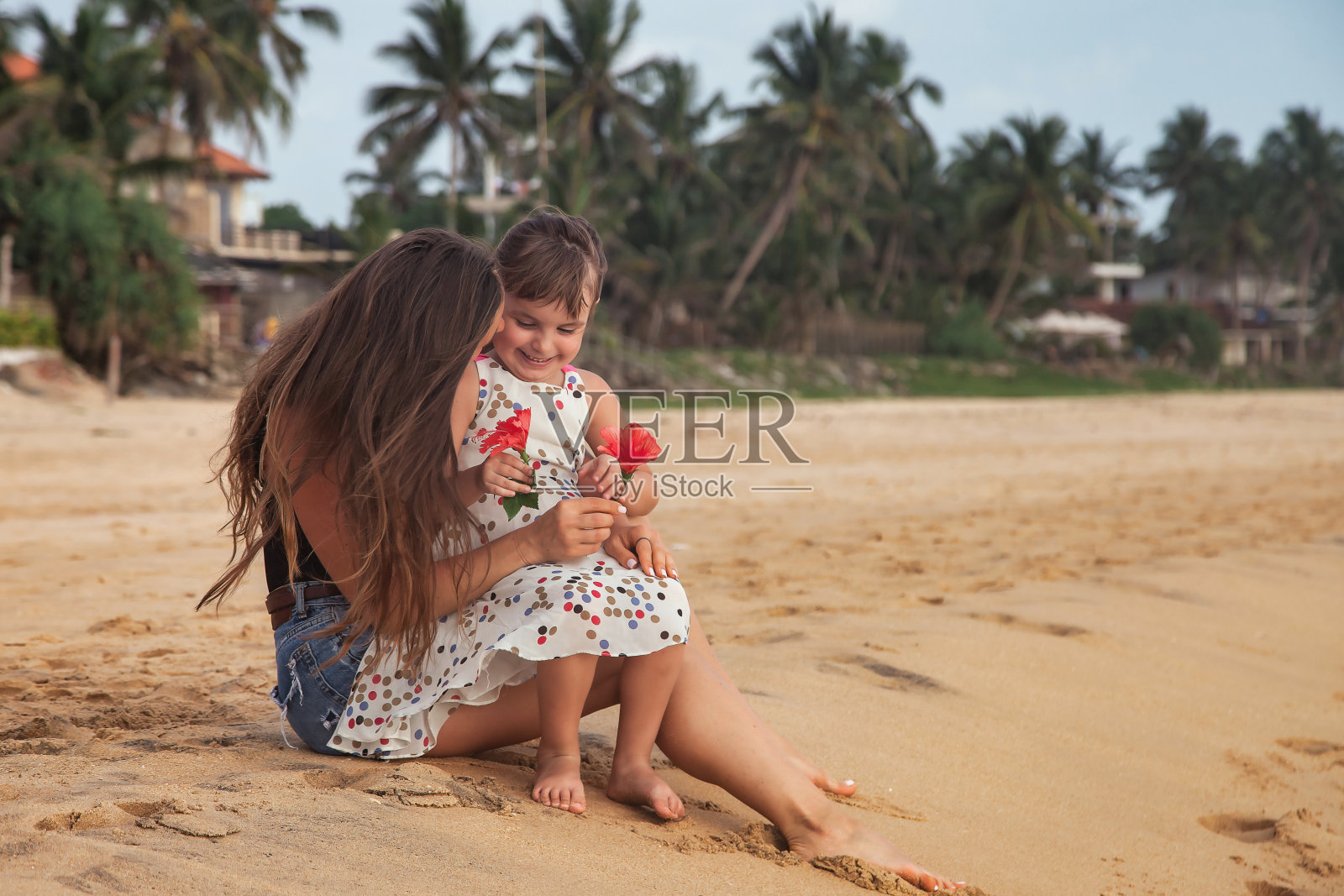 妈妈和小女儿一起坐在棕榈树背景下的热带沙滩上。母亲和小女孩穿着便装在海上。概念家庭度假，旅游。复制文本空间照片摄影图片
