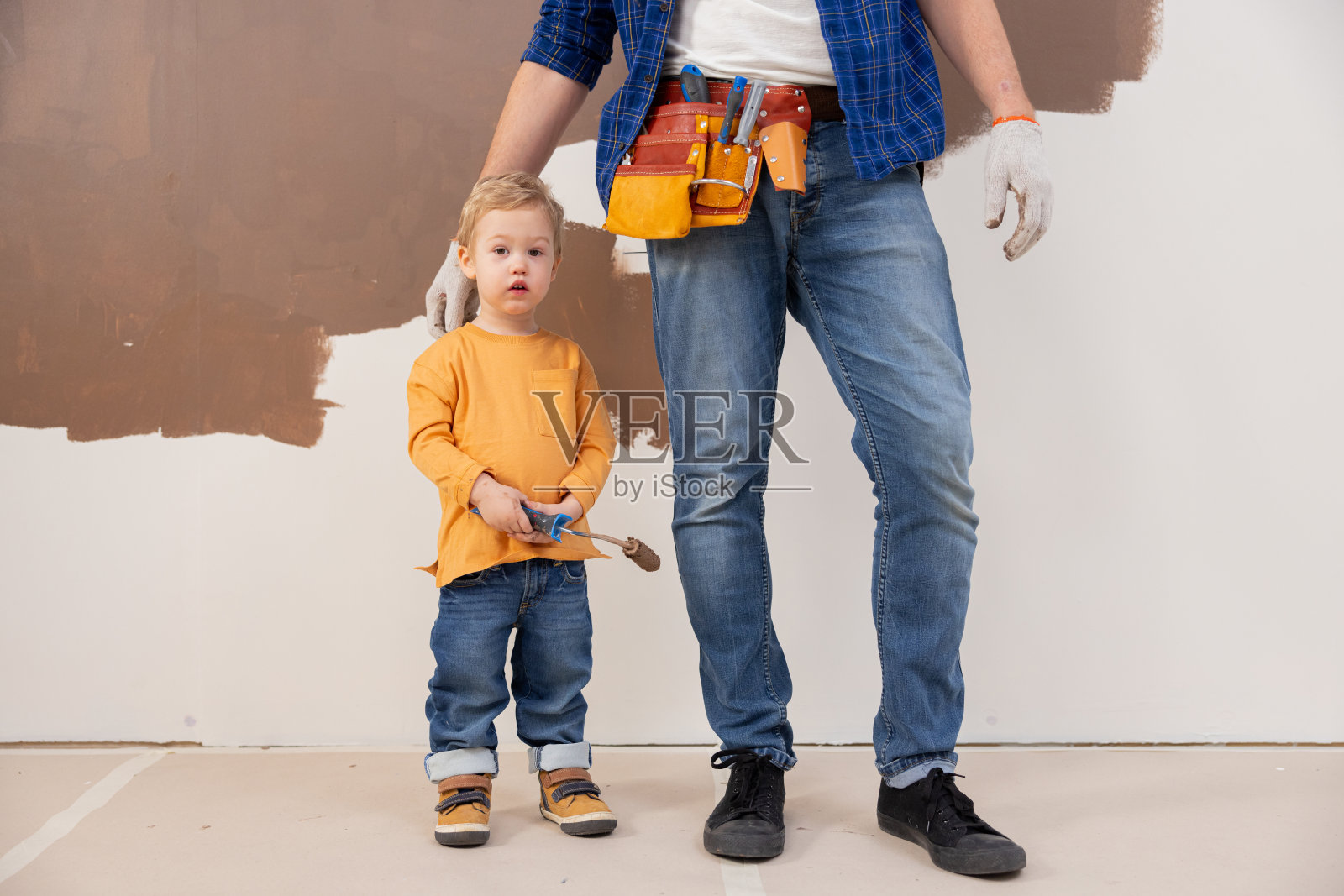 一个三岁的金发小男孩，站在他爸爸的脚边。那孩子手里拿着一个油漆滚筒。他的手沾满了油漆。对于一个孩子来说，这是第一次体验修复照片摄影图片