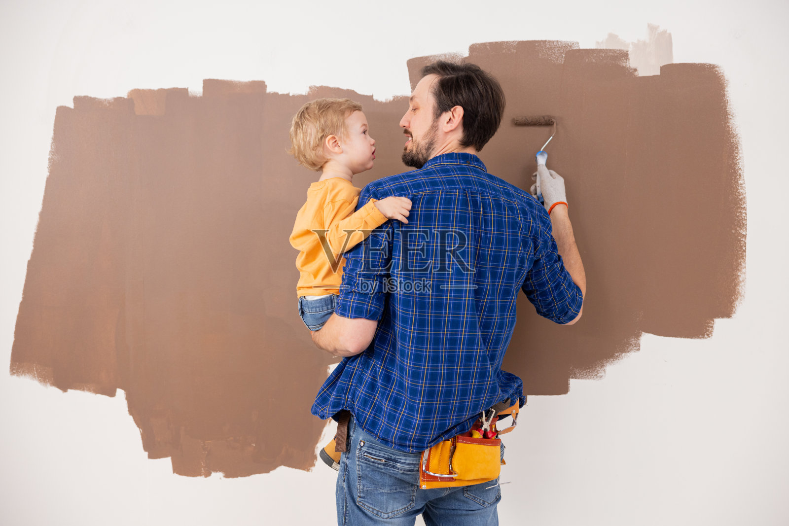 一位父亲抱着他的小儿子，他们一起画出了棕色的云朵。那个小孩穿着一件橙色的衬衫，那个男人穿着一件蓝色的格子衬衫。这张照片是从背面拍的照片摄影图片