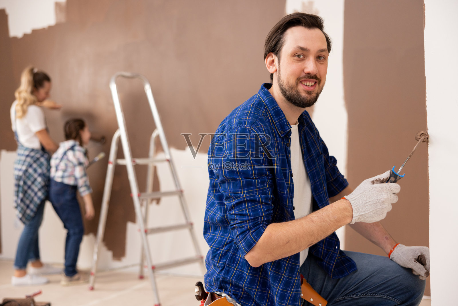 一个微笑着的黑发男人用一个小滚筒把墙涂成棕色。在他身后，从后面看，是他的妻子和女儿。他们都参与了维修工作照片摄影图片