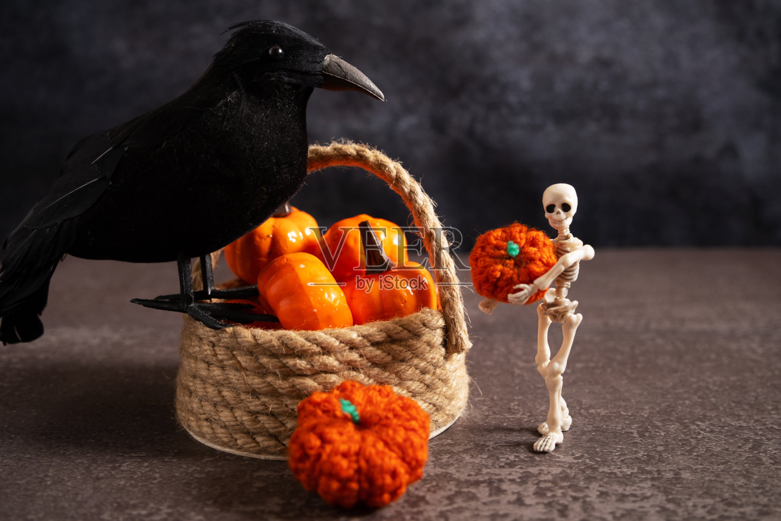 万圣节装饰安排柳条篮子，一个男人的骨架拿着一个南瓜，和一个黑乌鸦在灰色的桌子。照片摄影图片