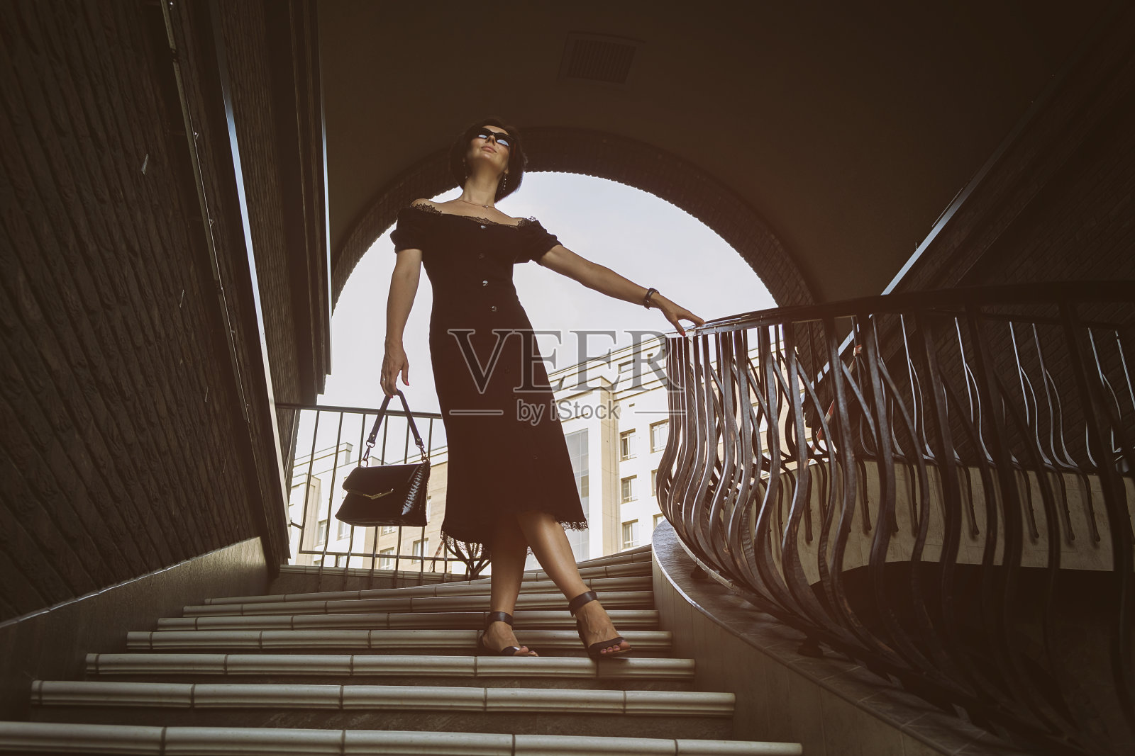 一个时髦的年轻女子，戴着帽子，太阳镜和黑色连衣裙走下街道的楼梯在一个夏天照片摄影图片