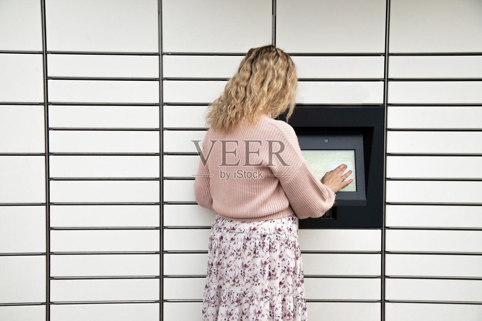 背面的年轻女子波浪金发在裙子站在米色自助送货柜，看着屏幕。照片摄影图片