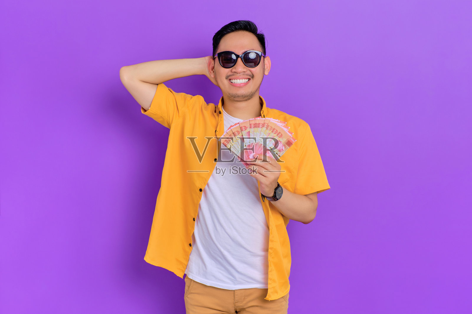 快乐英俊的年轻亚洲男子戴着太阳镜，拿着纸钱，庆祝大运气孤立在紫色的背景照片摄影图片
