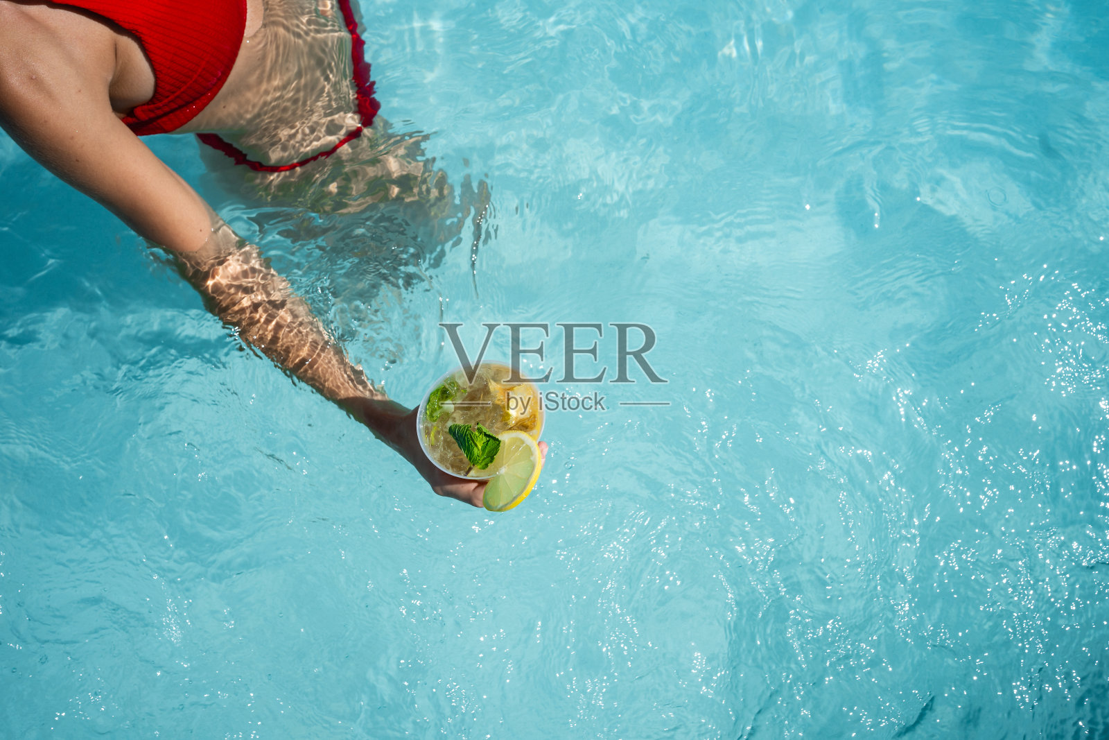 夏天，身穿红色比基尼的女子在游泳池里喝着莫吉托鸡尾酒照片摄影图片