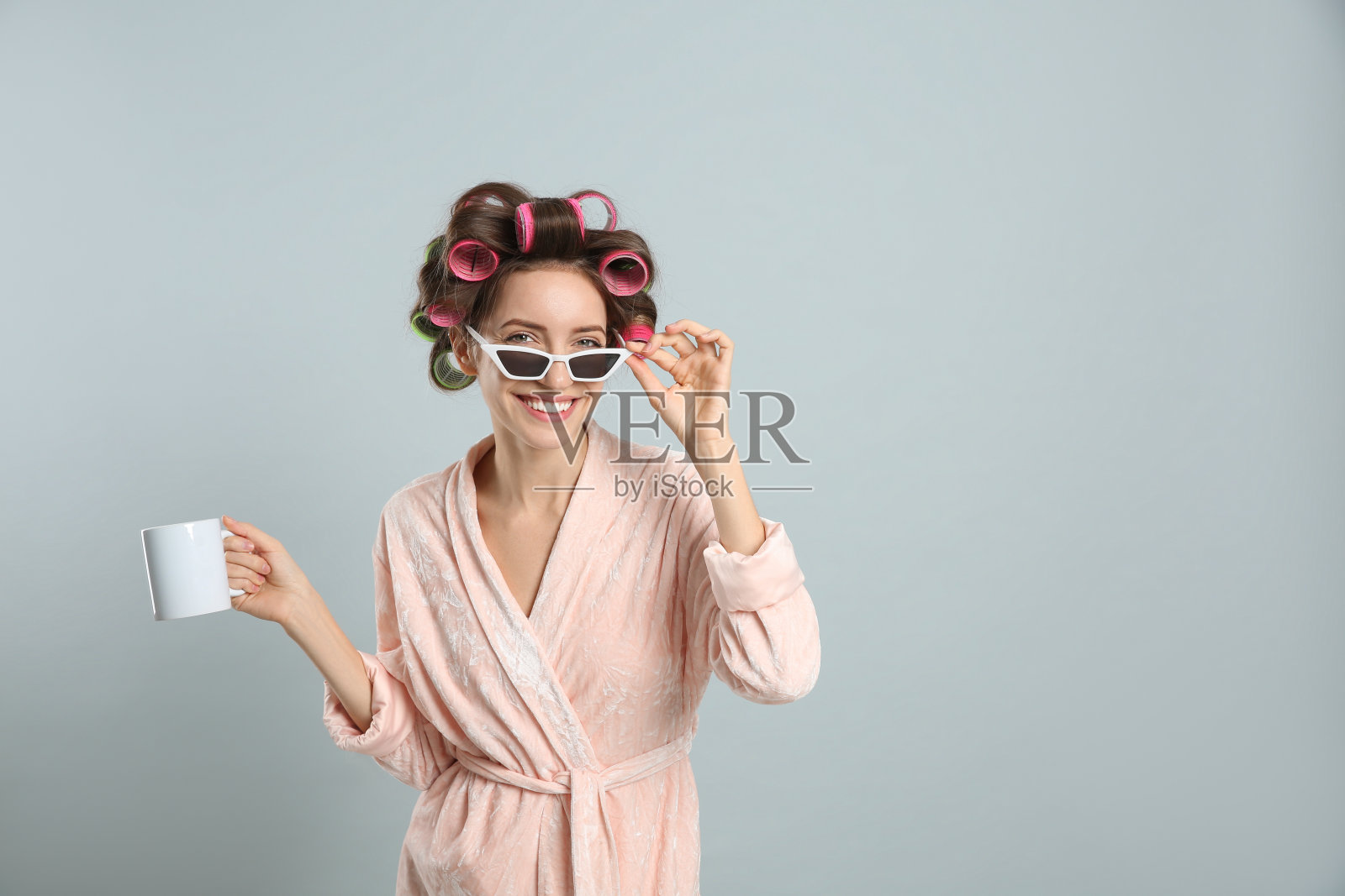 美丽的年轻女子在浴袍与卷发手拿杯饮料浅灰色的背景。空间的文本照片摄影图片
